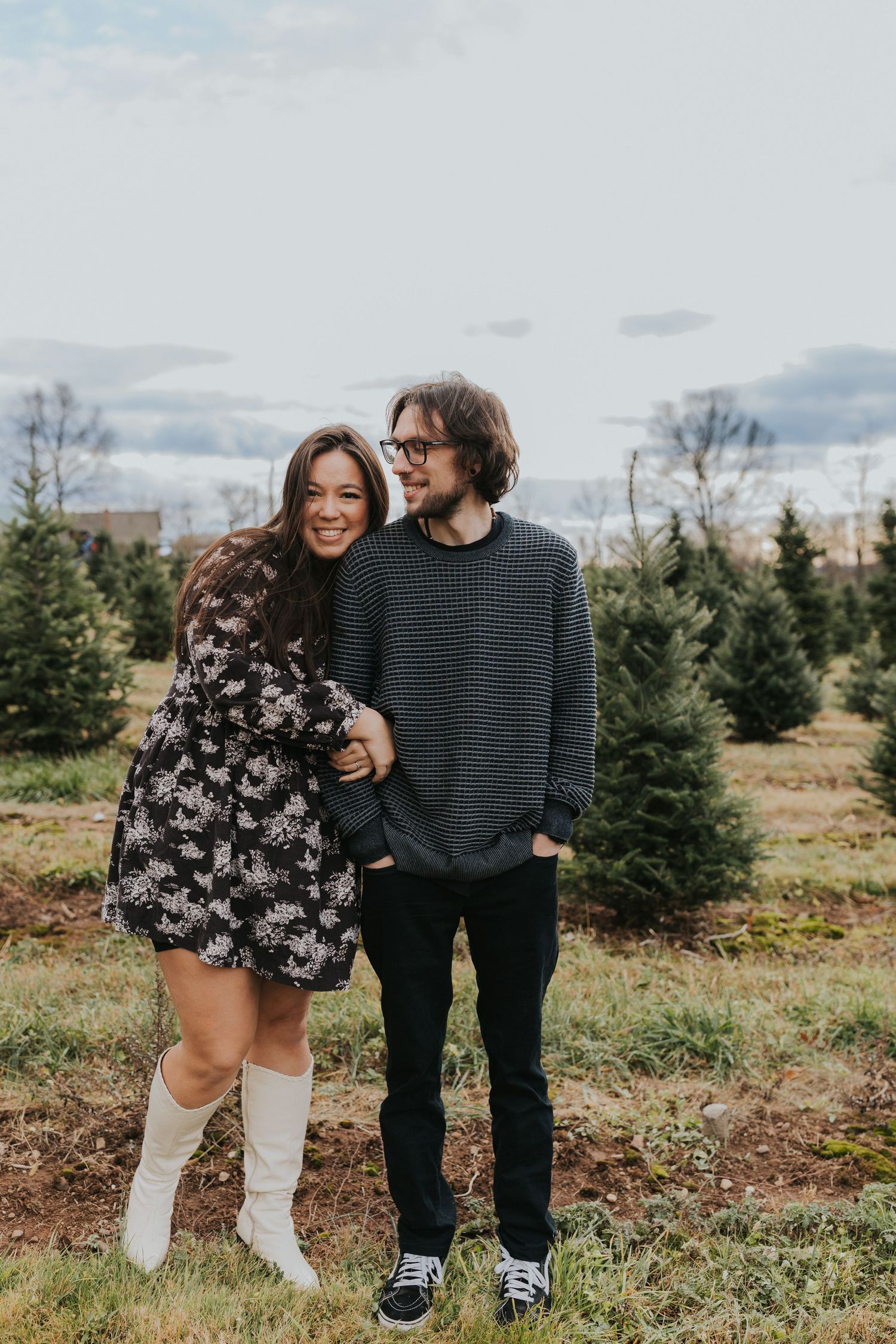 Smiling couple embracing arm in arm at a Christmas tree farm on a cloudy day.