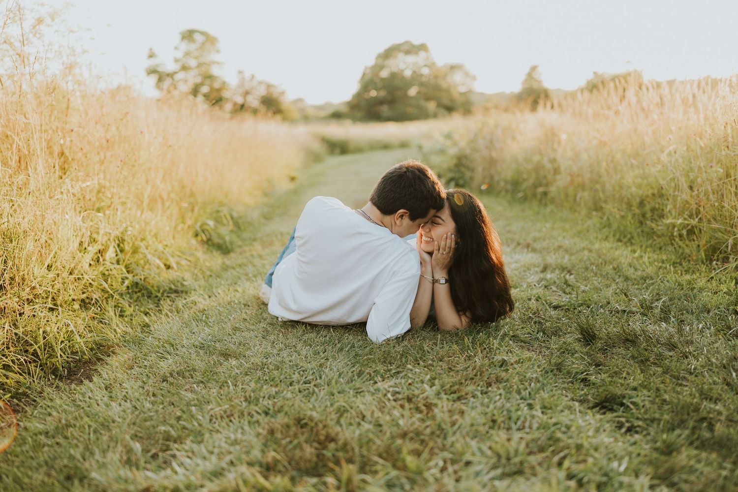 Couple lying in a grassy field path surrounded by golden meadows during a romantic outdoor engagement session.