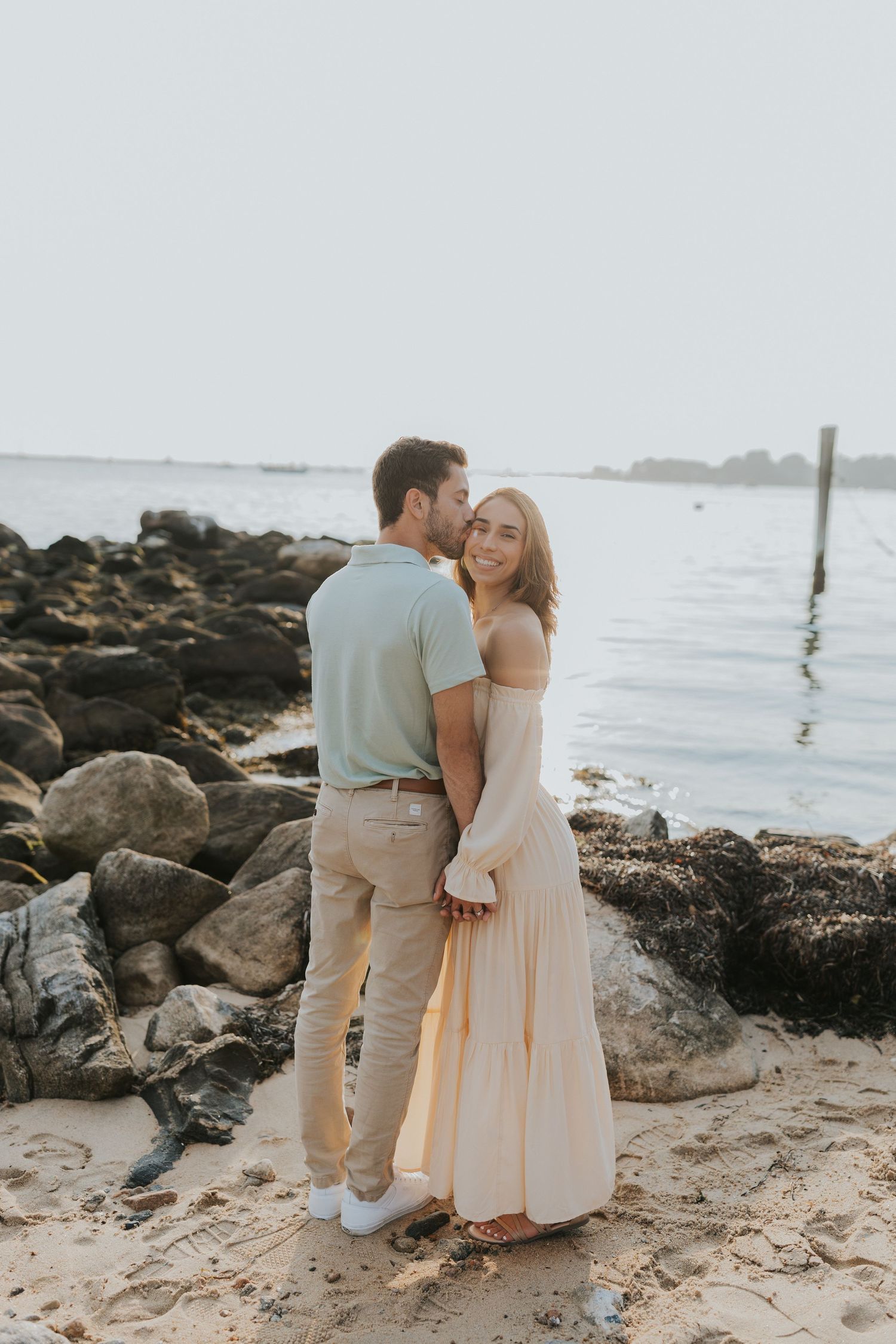 Romantic couple embracing on a rocky beach at dusk, woman in flowing cream maxi dress smiling at camera.