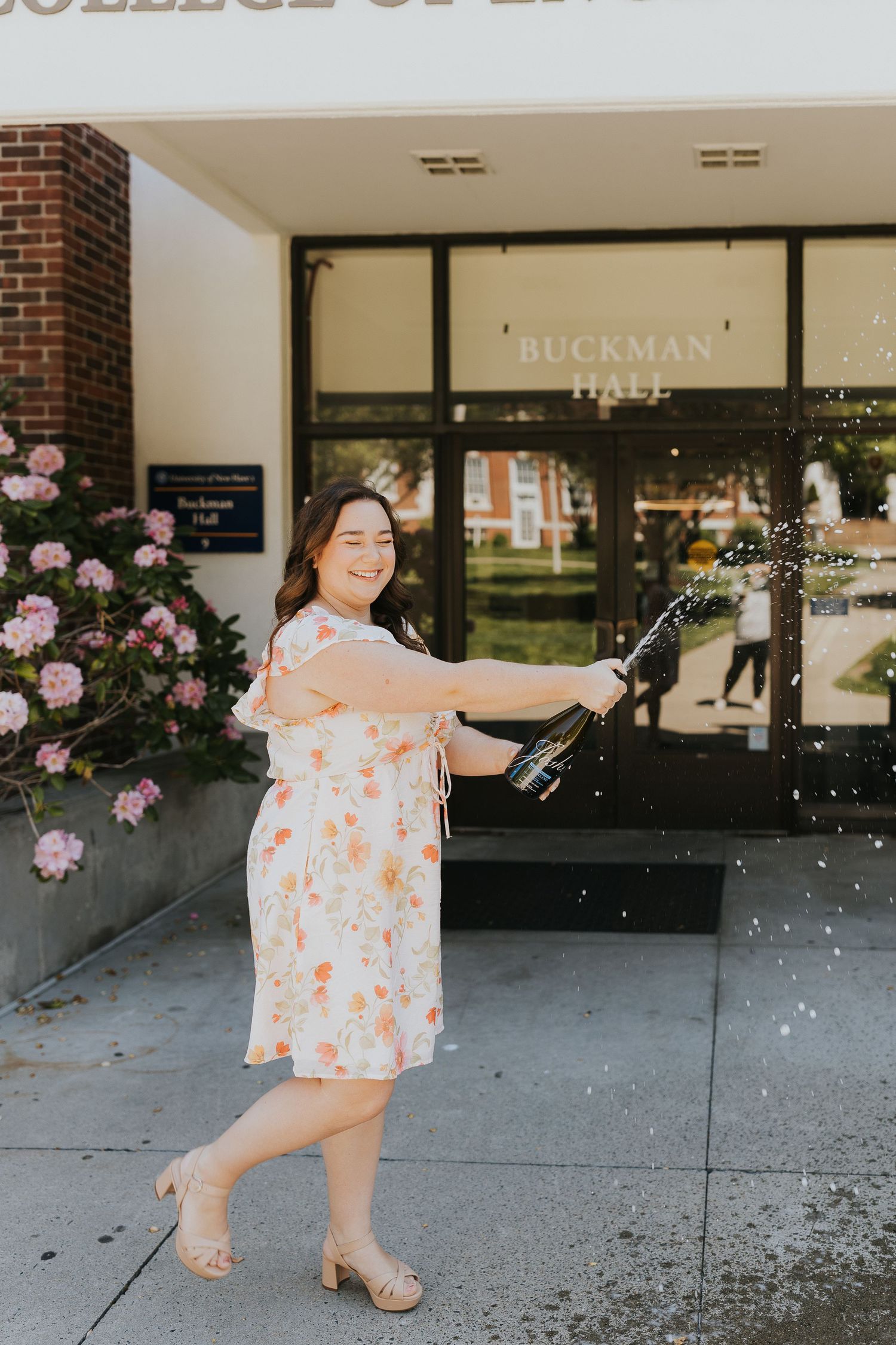 Graduate in floral dress pops champagne bottle outside Buckman Hall entrance.