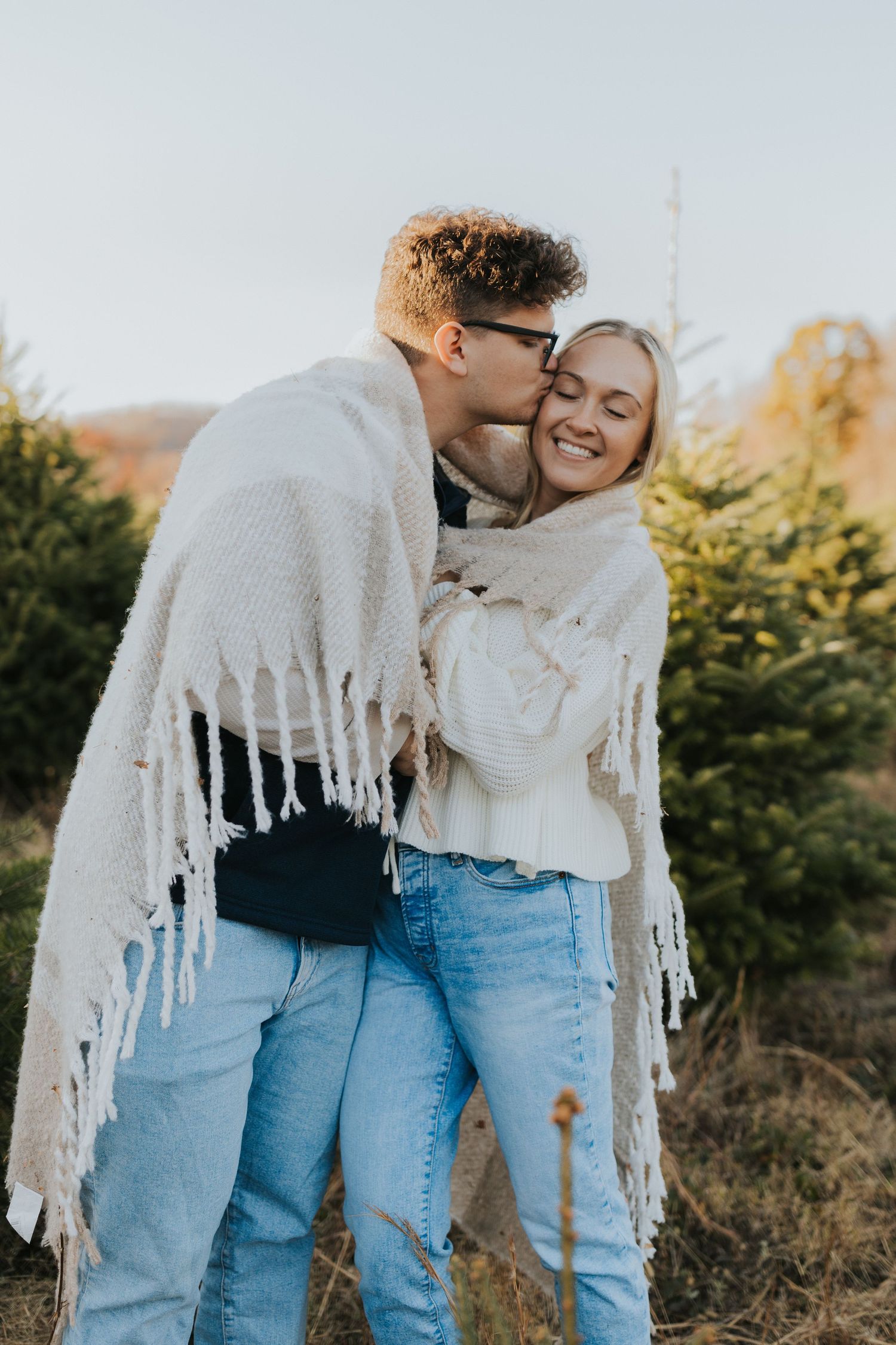 Couple wrapped in a white fringe blanket sharing a kiss at a Christmas tree farm at sunset.