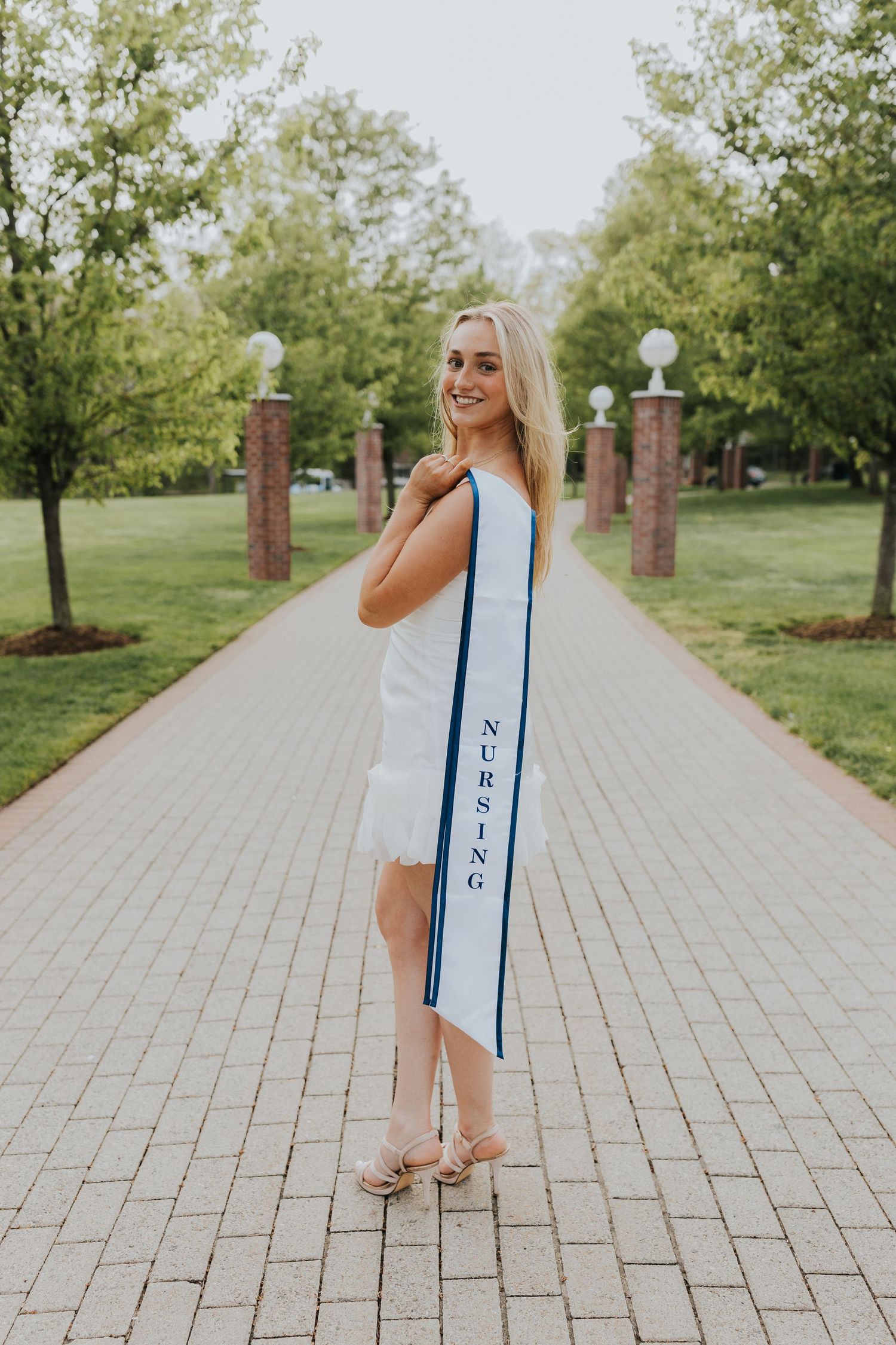 Nursing graduate in white dress holding long stole on a scenic brick campus pathway.