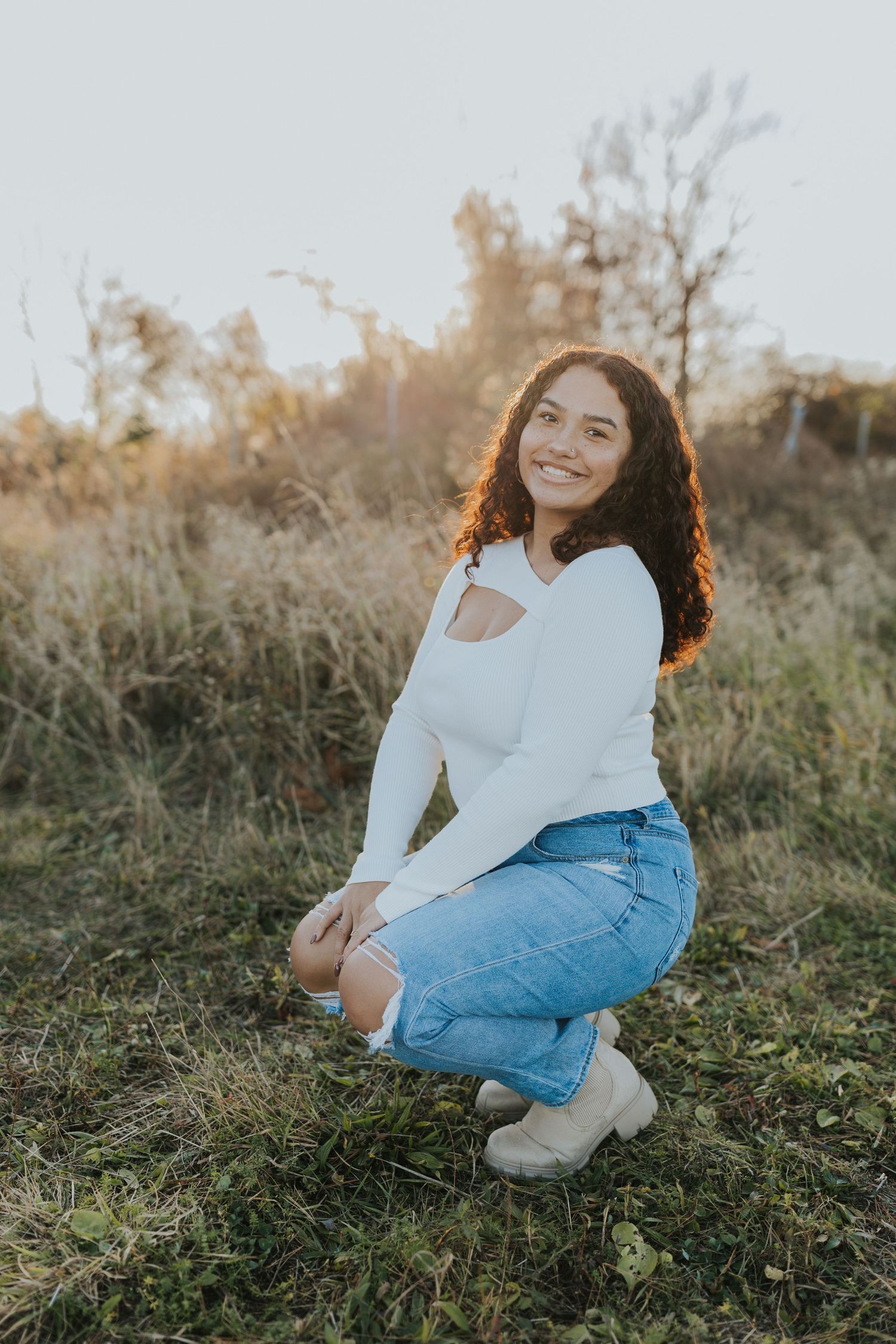 Smiling young woman with curly hair crouching in white top and denim skirt in autumn field.