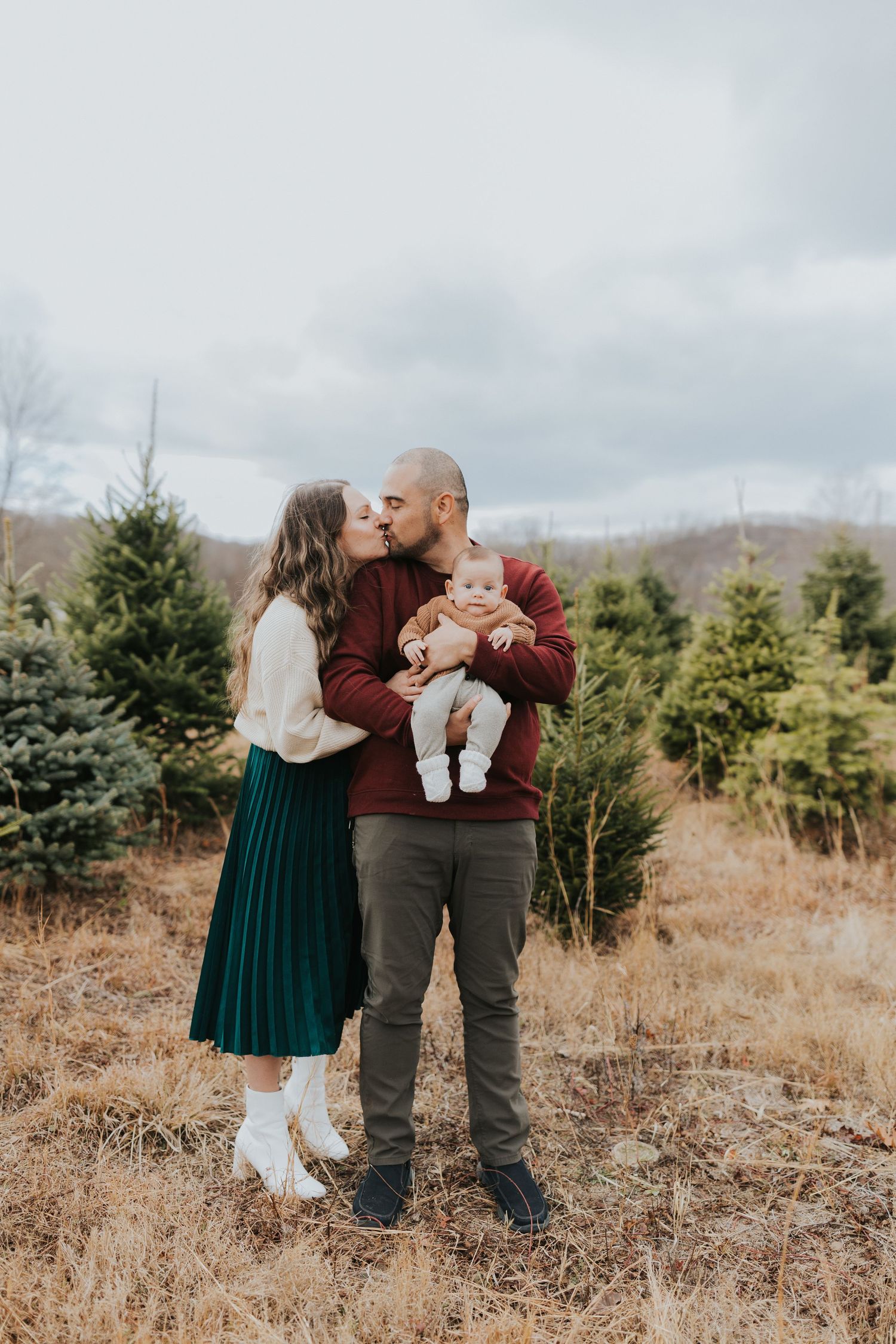 Parents sharing a kiss while father holds baby at a Christmas tree farm outdoor setting.