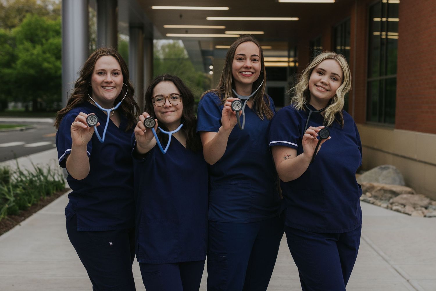 Four nursing students in navy scrubs proudly display their stethoscopes outside a medical building.
