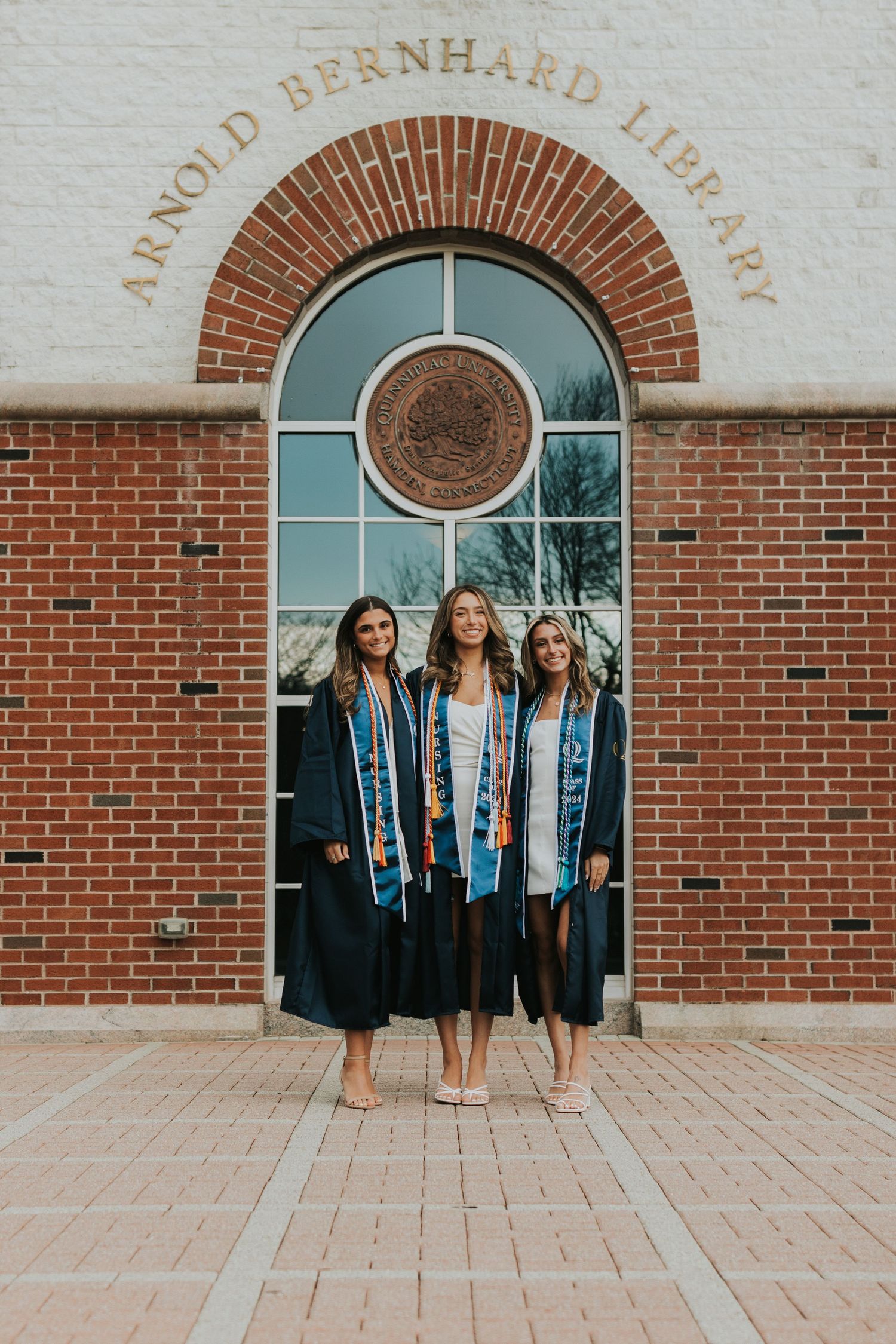 Three graduates in navy nursing gowns and stoles pose together at Arnold Bernhard Library entrance.