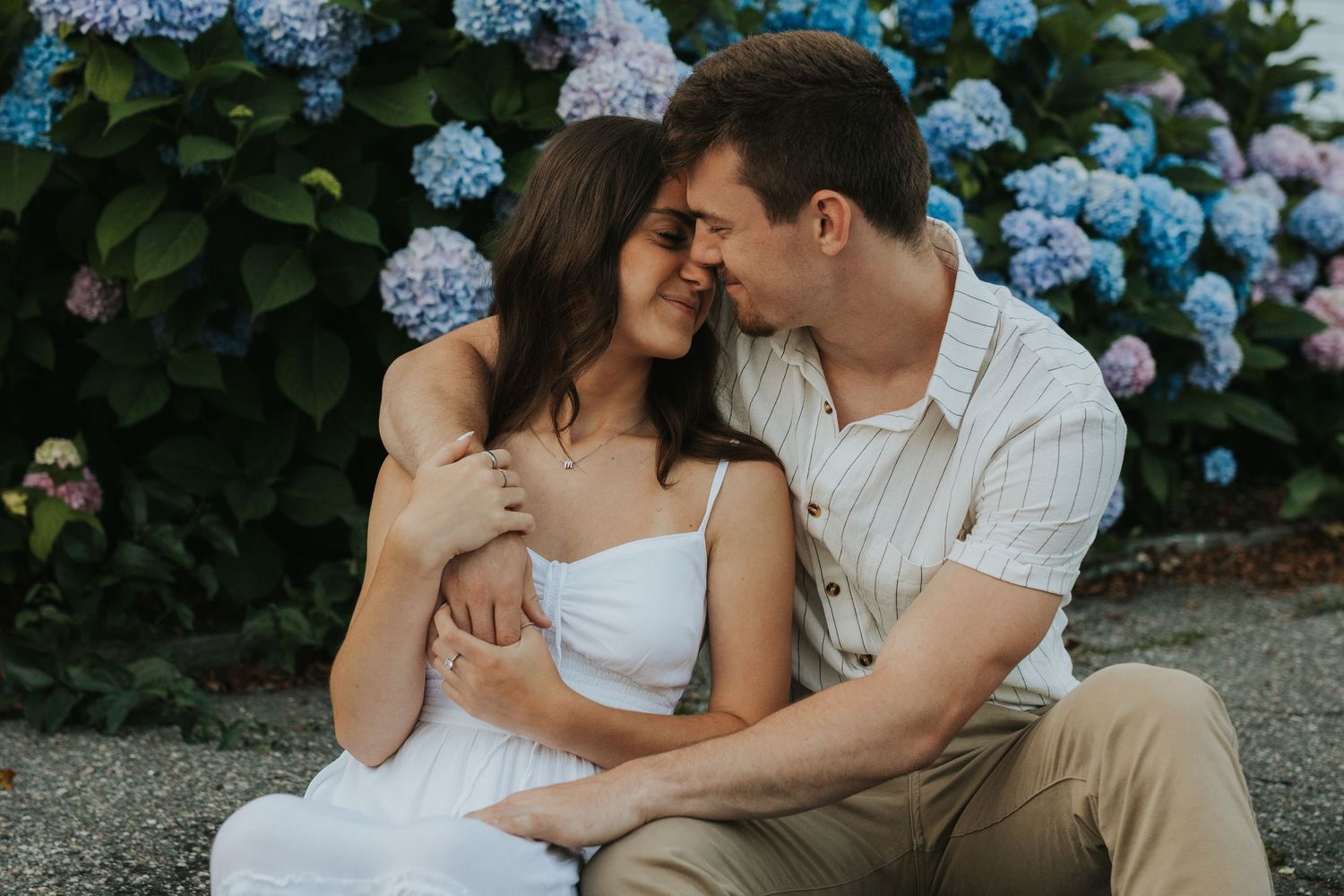 Romantic couple sitting together nose-to-nose in front of blooming blue hydrangea bushes outdoors.