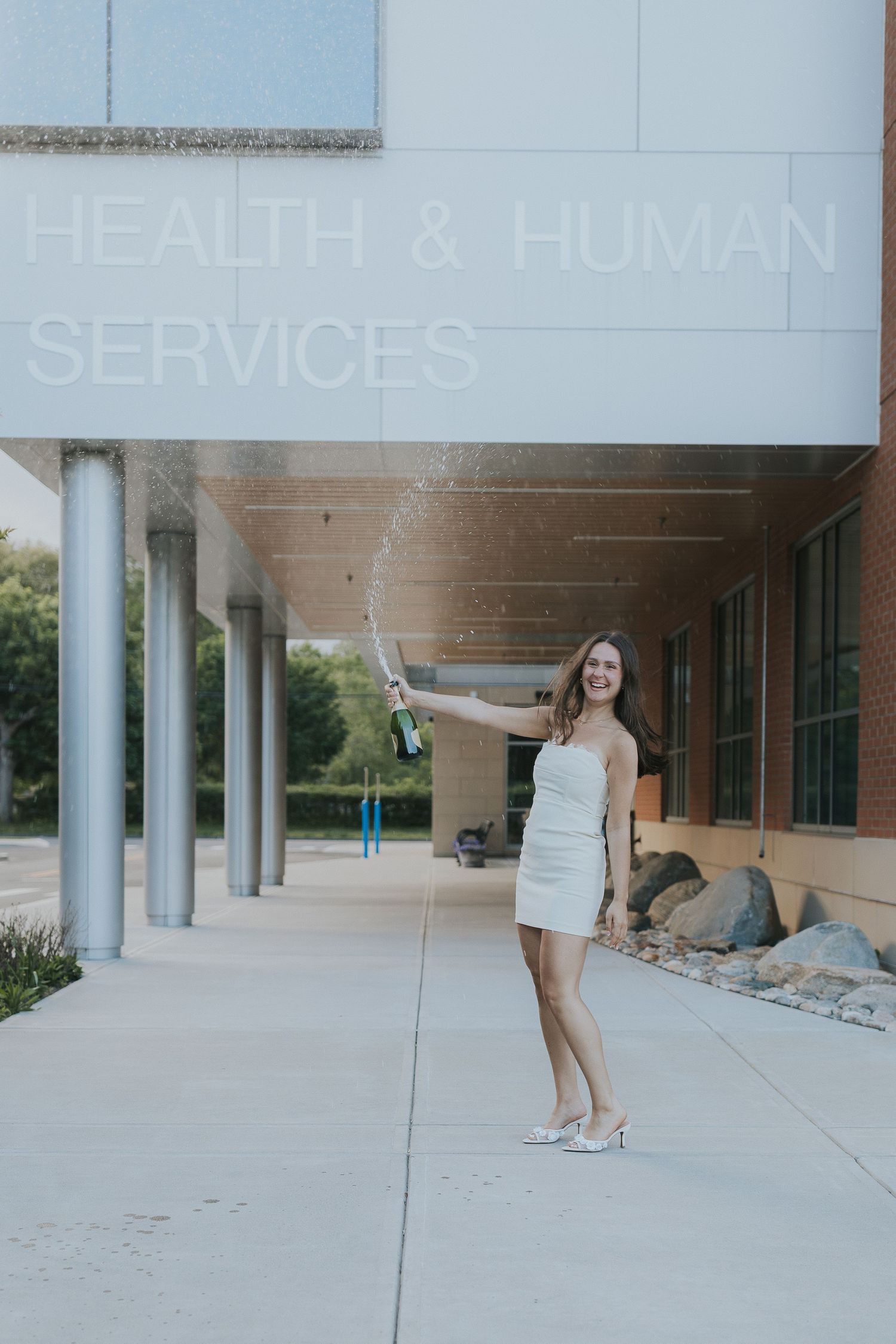 Woman in white dress spraying champagne outside Health & Human Services building, celebrating graduation.