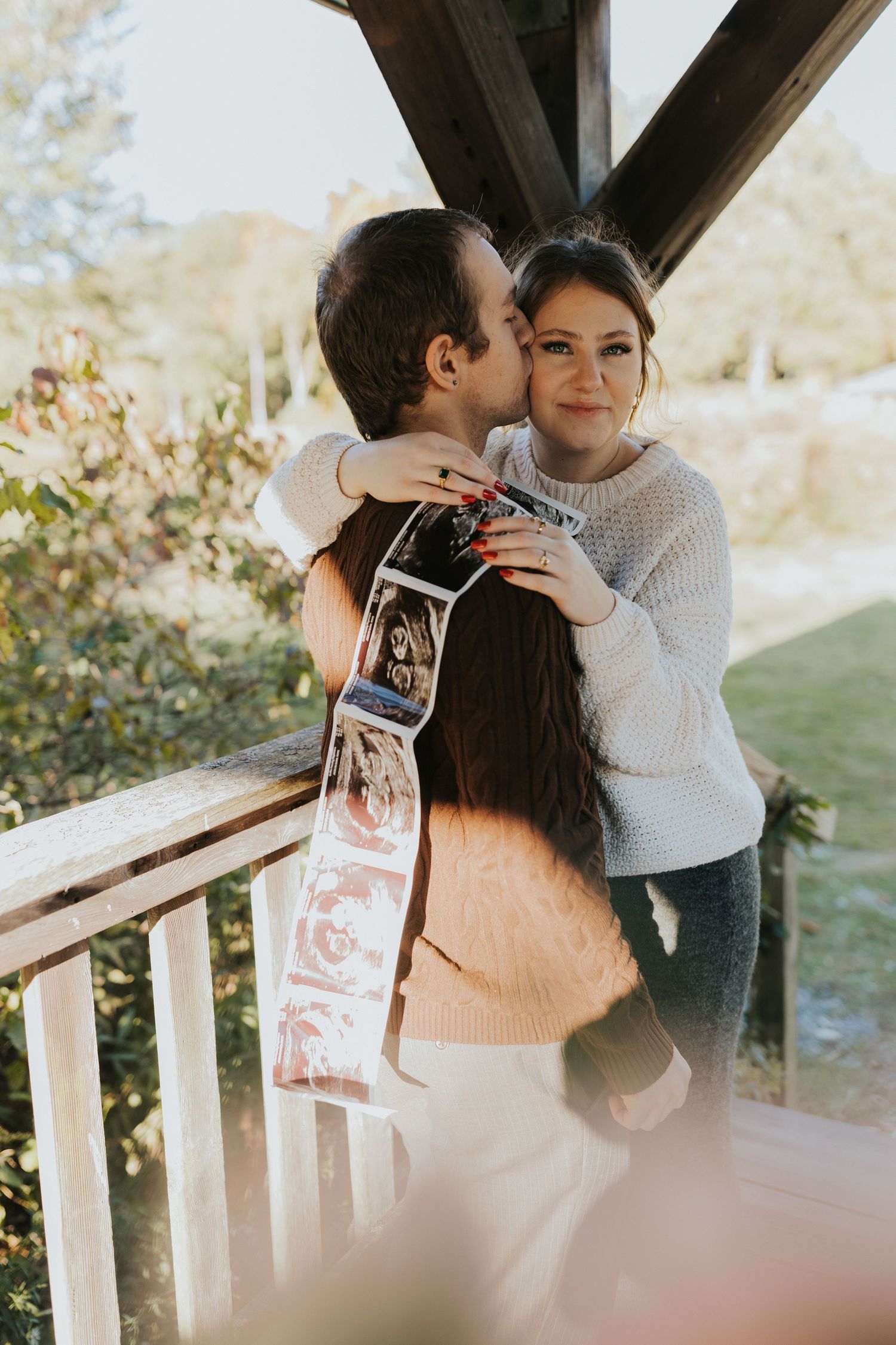 Couple sharing a kiss on wooden porch, woman holding ultrasound photos for pregnancy announcement.