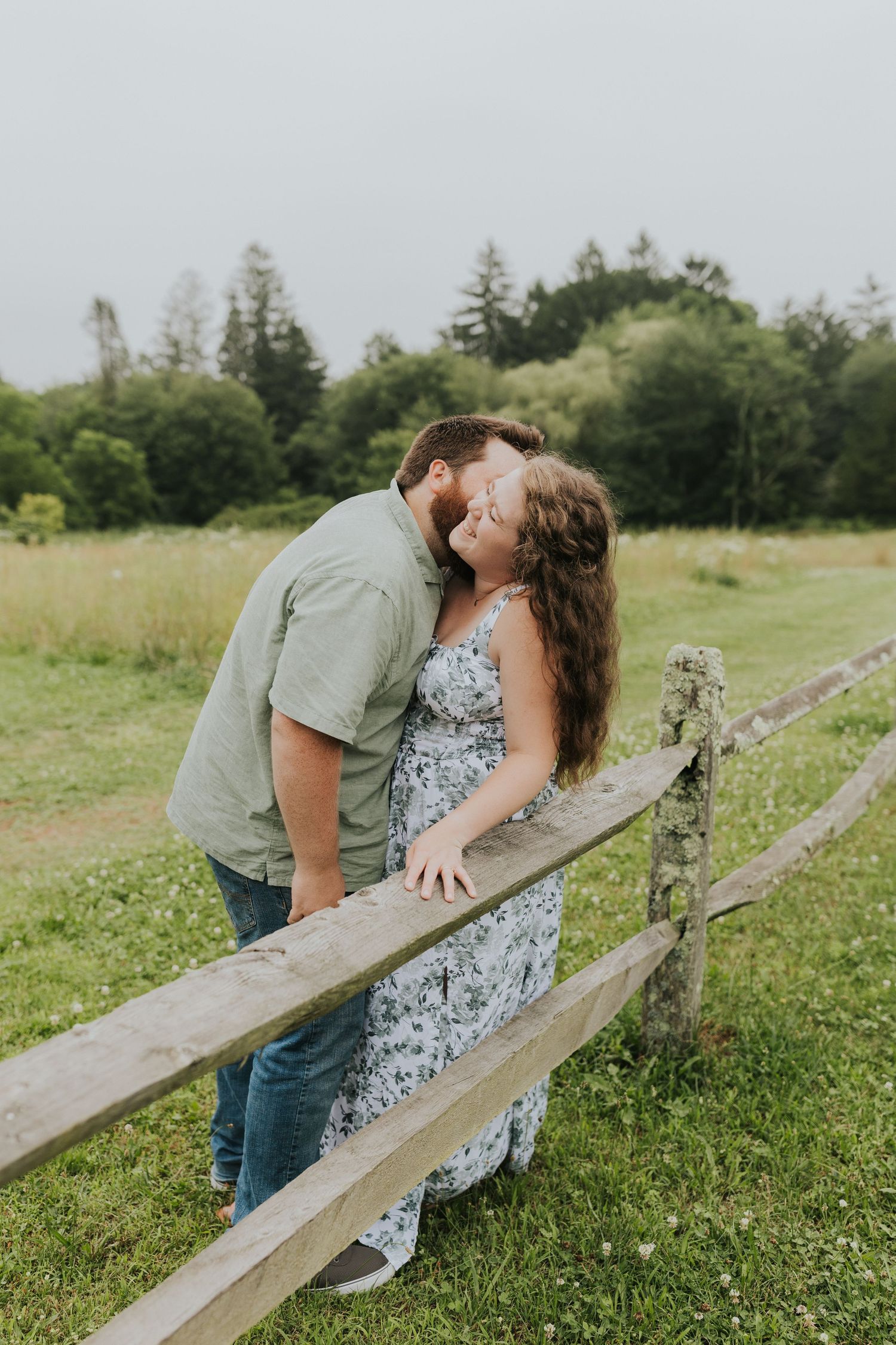 Couple sharing a sweet kiss near a rustic wooden fence in a green countryside meadow setting.