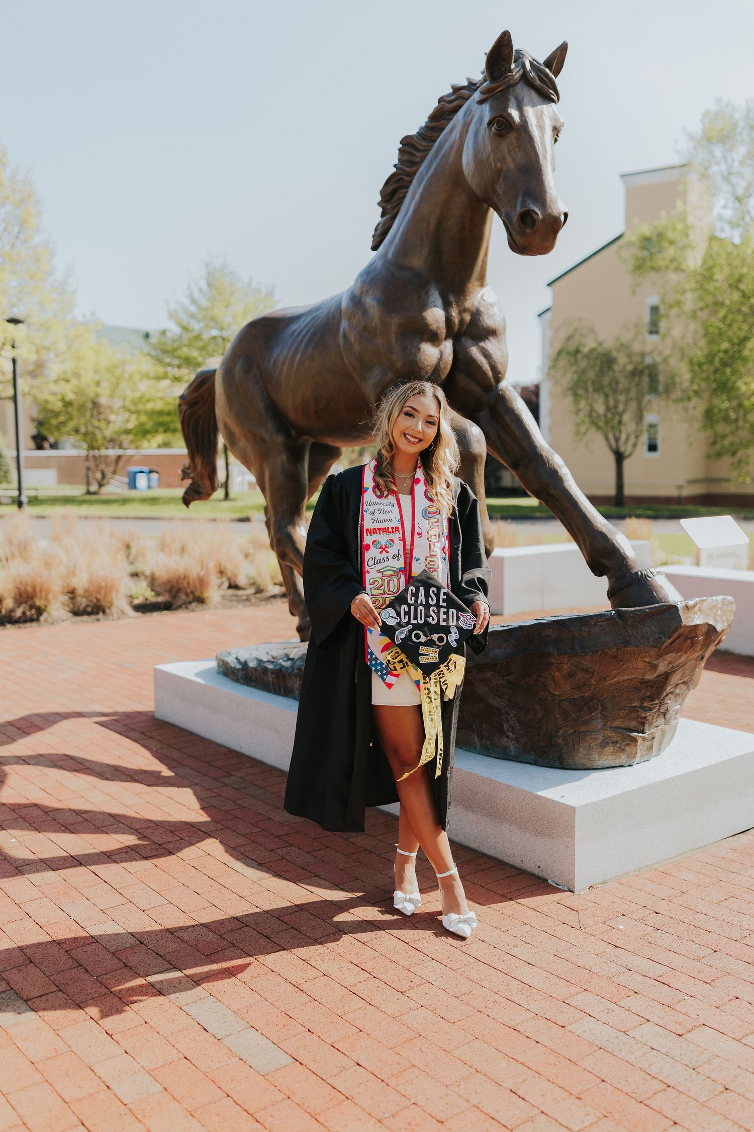 Smiling graduate in black gown poses with Case Closed cap beside bronze horse statue on brick university plaza.