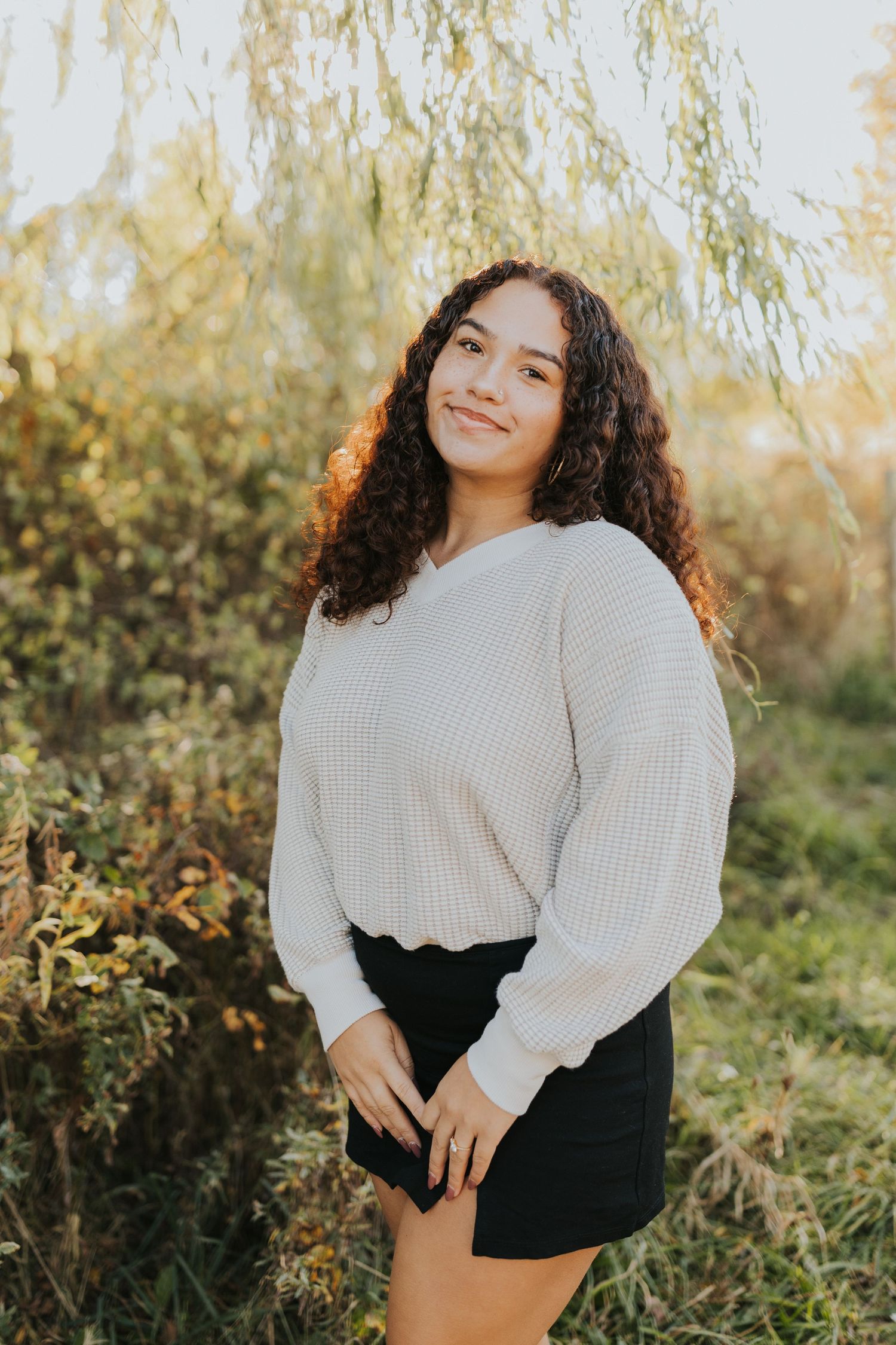 Young woman with curly hair in cream sweater and black skirt smiling outdoors at sunset.