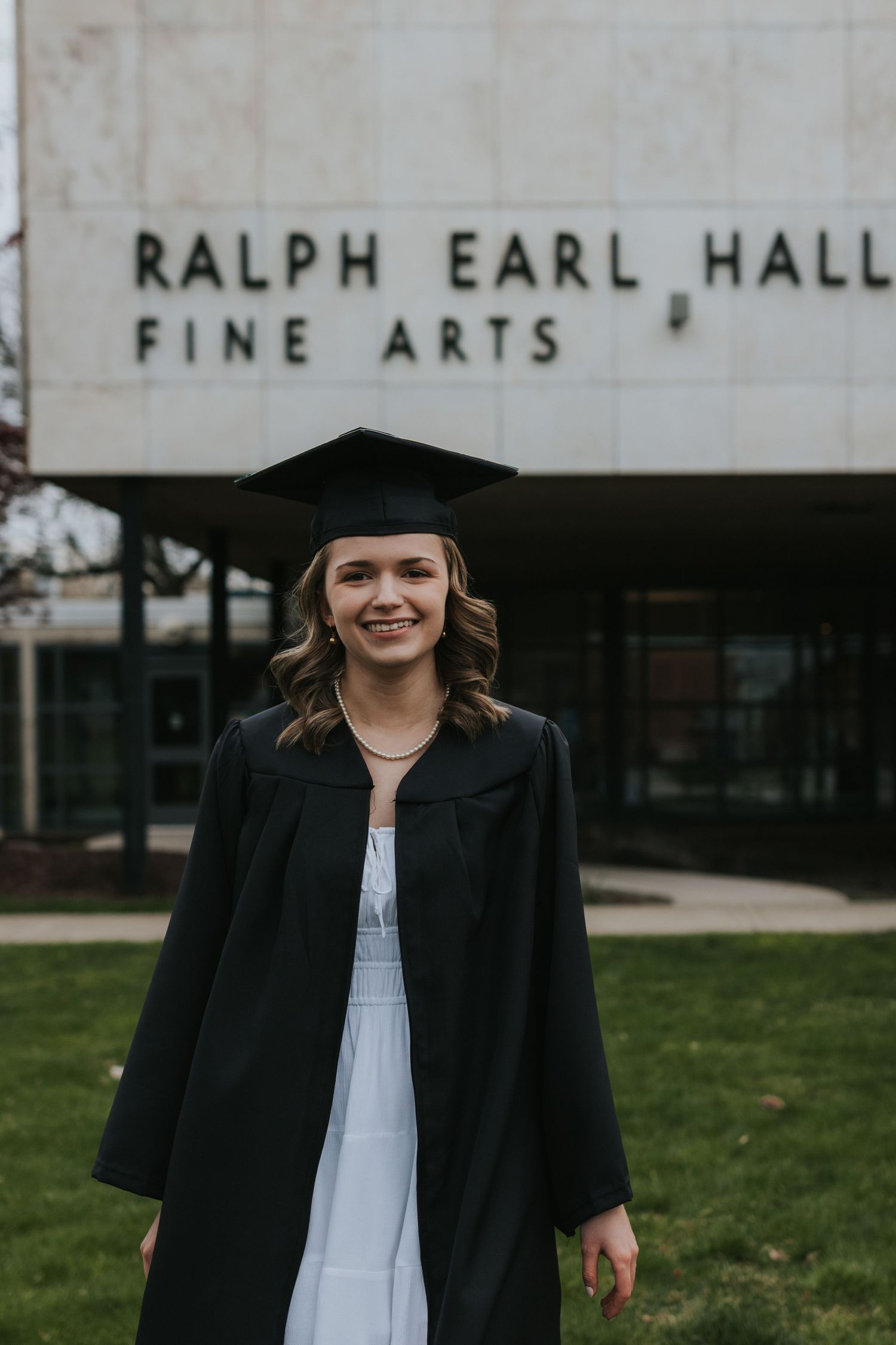 Female graduate in cap and gown smiles outside Ralph Earl Hall Fine Arts building.