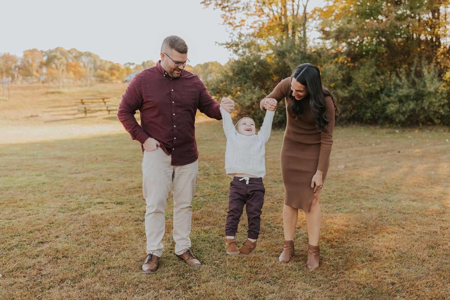 Parents swinging toddler son by hands in open field during colorful autumn family photo session.