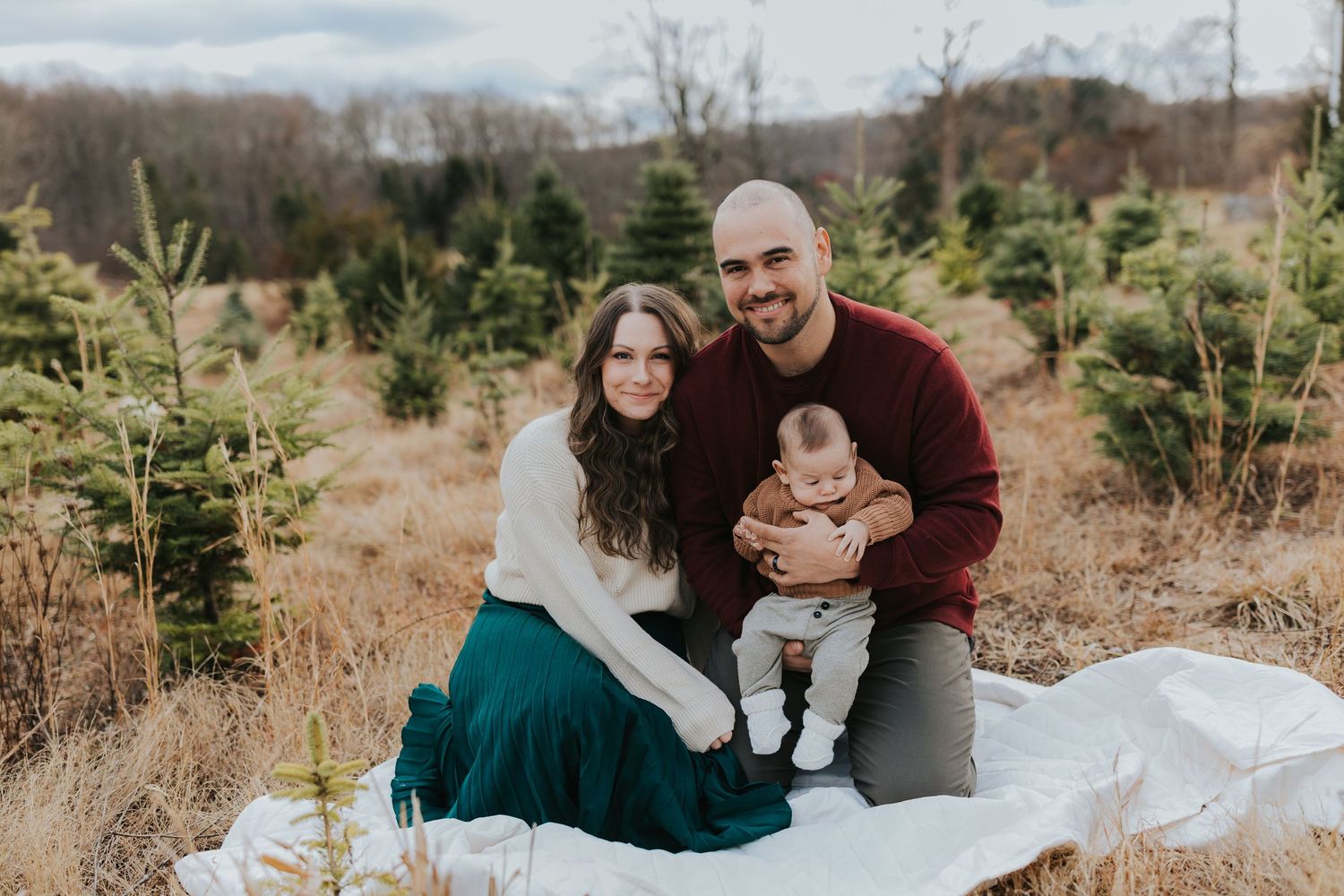 Family portrait at Christmas tree farm, parents sitting with infant on white blanket outdoors.