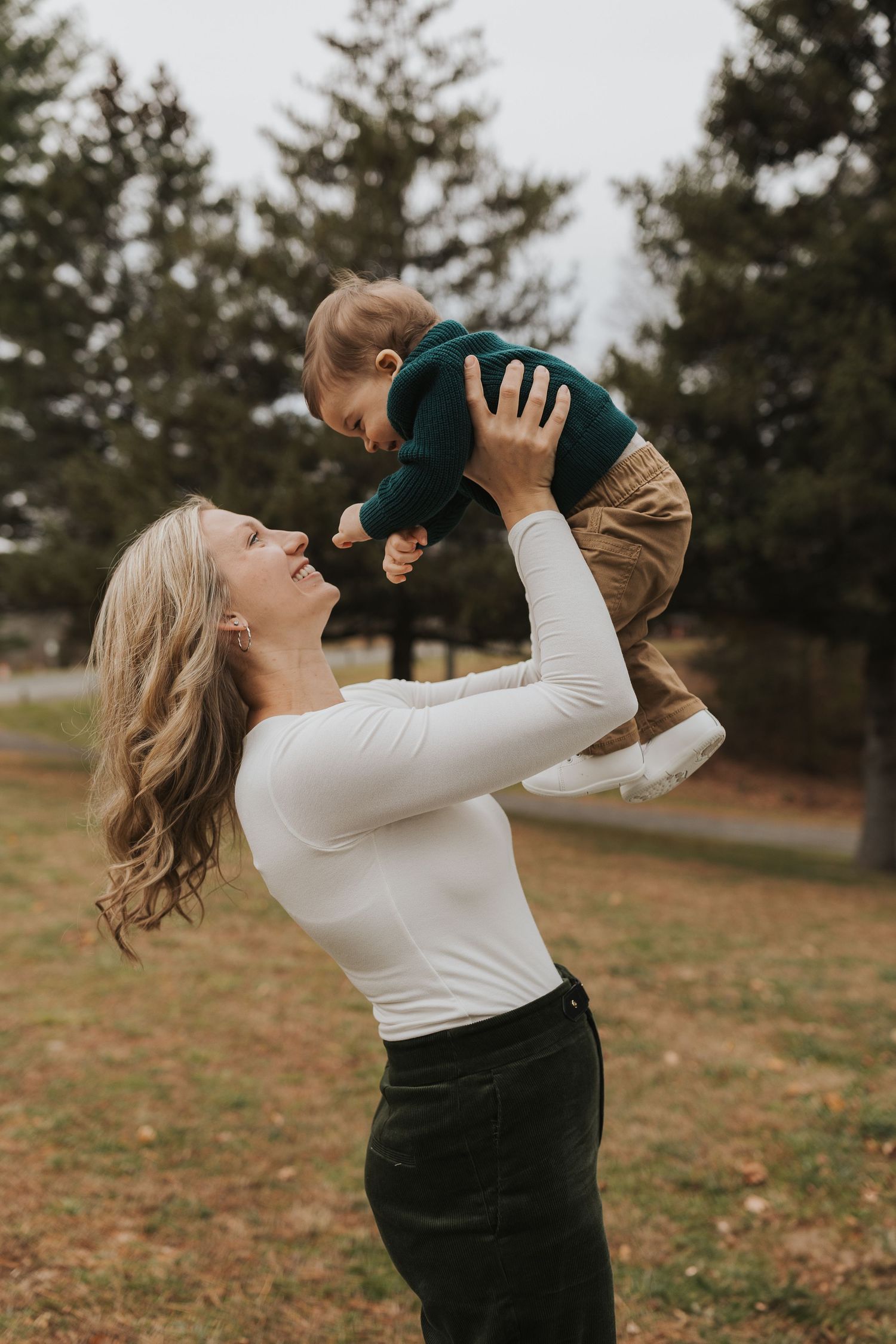 Joyful blonde mother lifting her baby boy in a green outfit in an autumn park.
