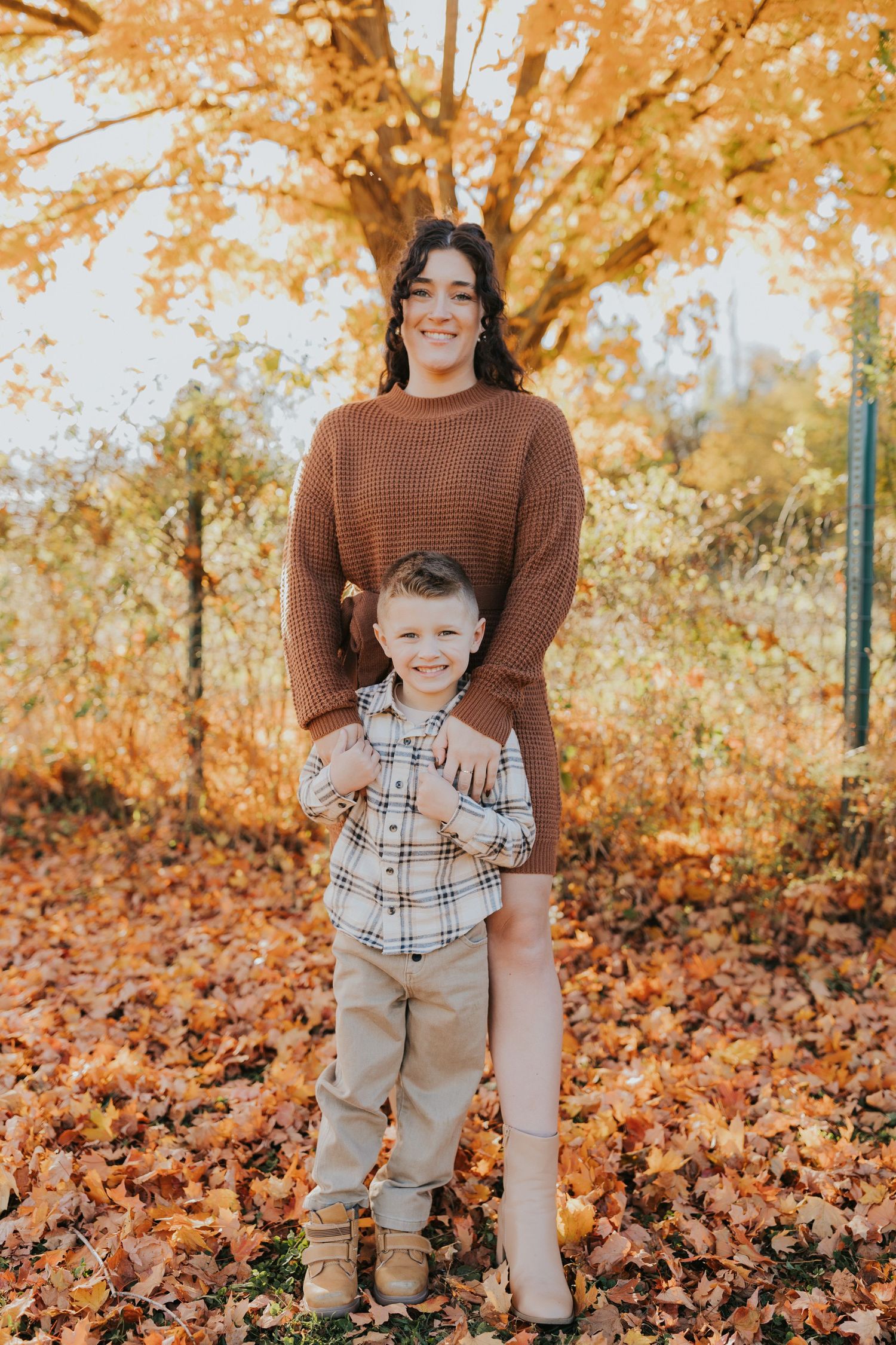 A mother and young son pose together among vibrant orange fall leaves beneath a golden tree.