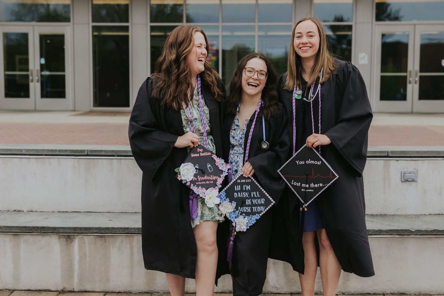 Three laughing graduates in black gowns displaying decorated caps outdoors.