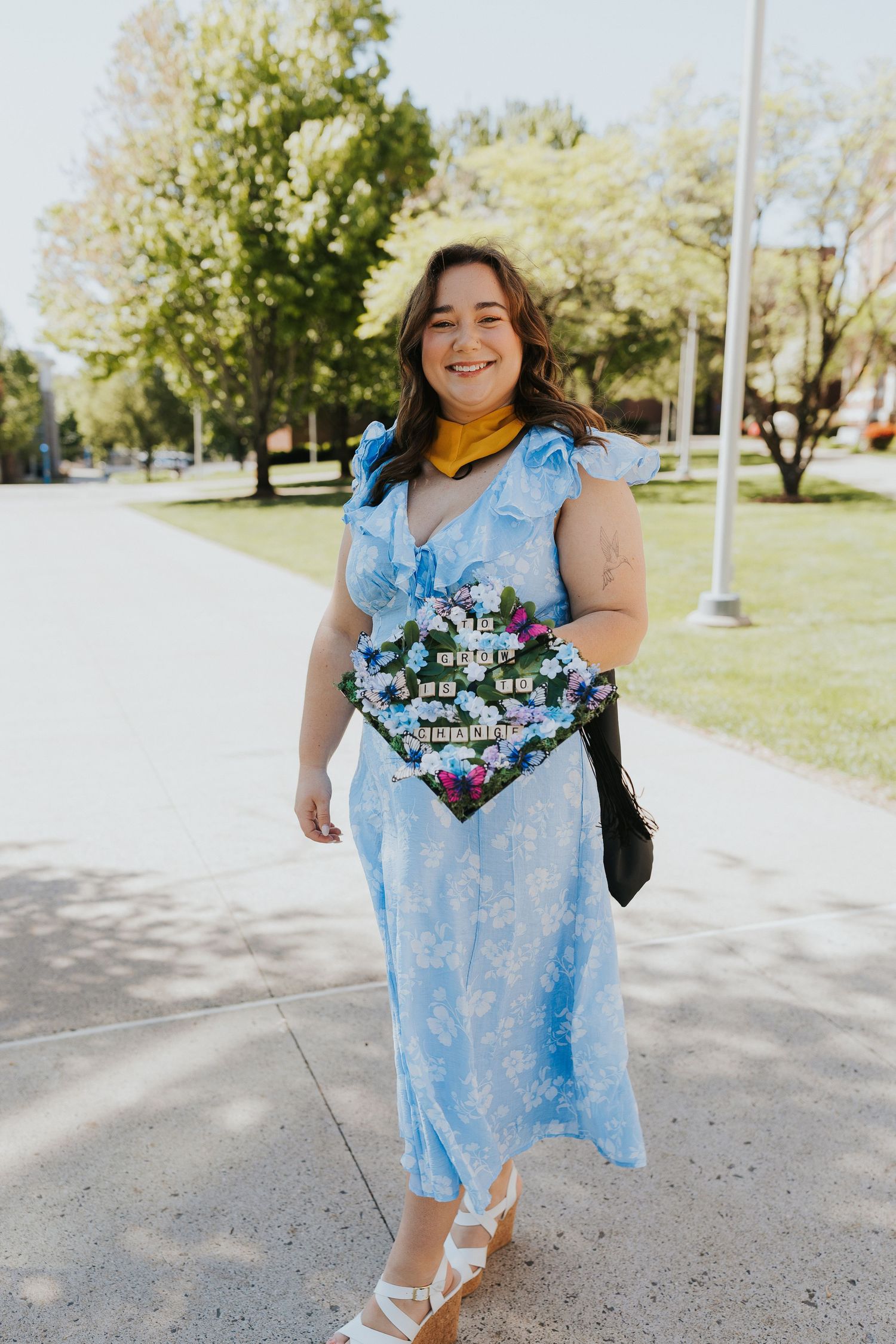 Smiling graduate in blue floral dress holding decorated cap with flowers on a sunny campus path.
