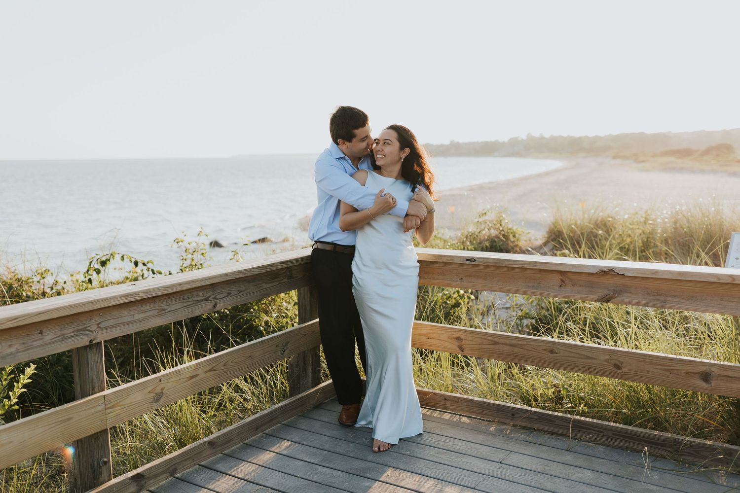 Couple embracing on a wooden beach boardwalk at golden hour, with ocean and sandy shore in the background.
