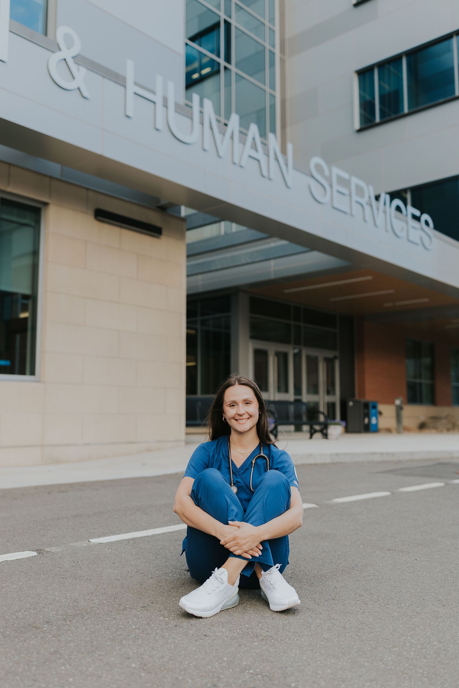 Female nursing student in blue scrubs sitting on ground outside Human Services building smiling.