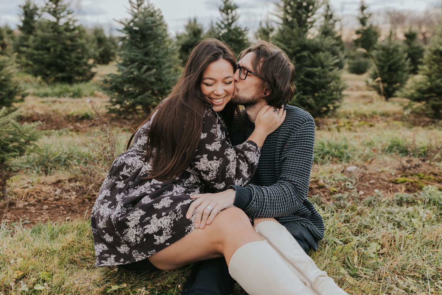 Two women share a tender kiss at a Christmas tree farm during a fall engagement photo session.