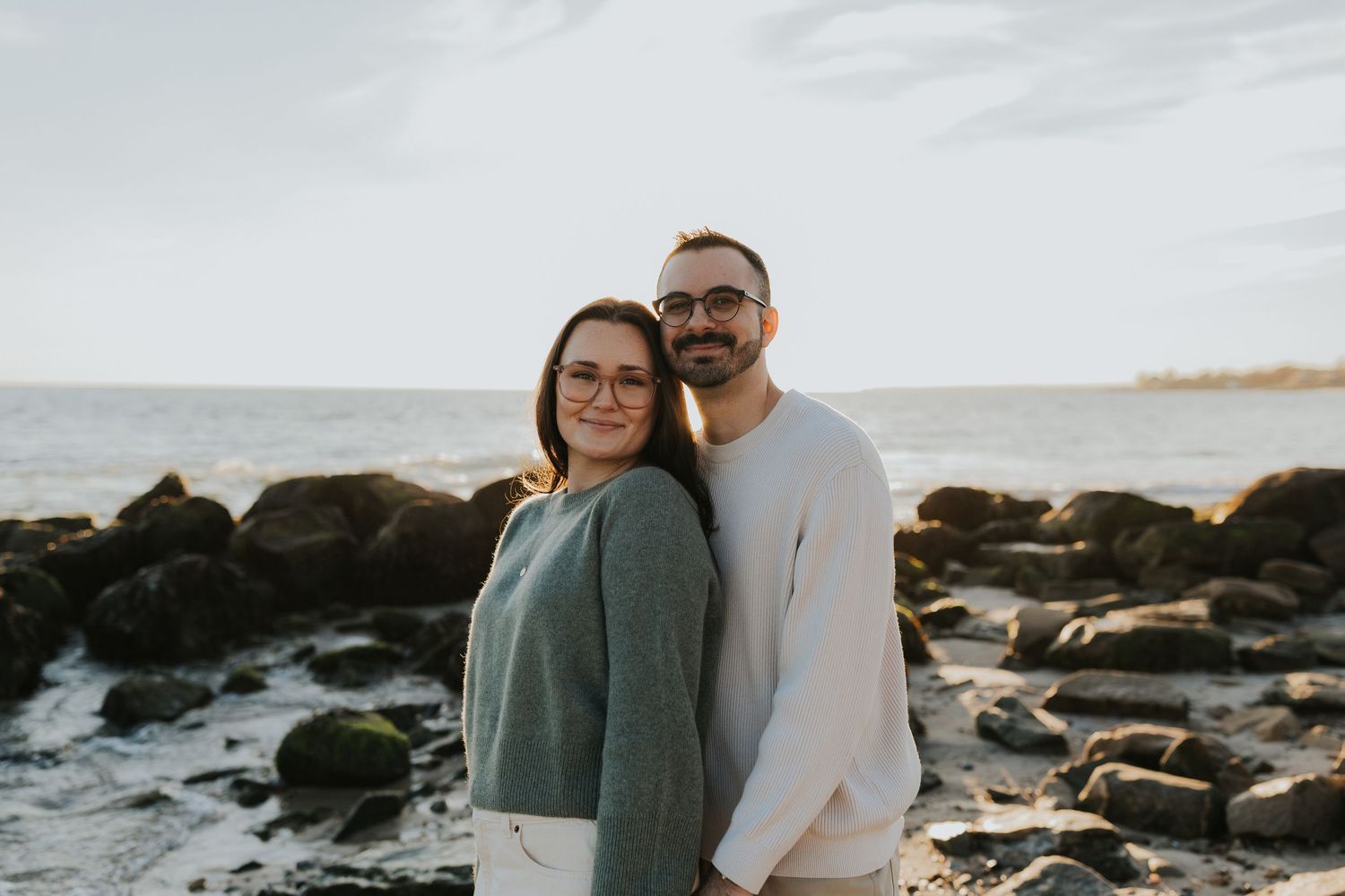 Couple standing together on rocky ocean shore at sunset, wearing matching green and cream sweaters.