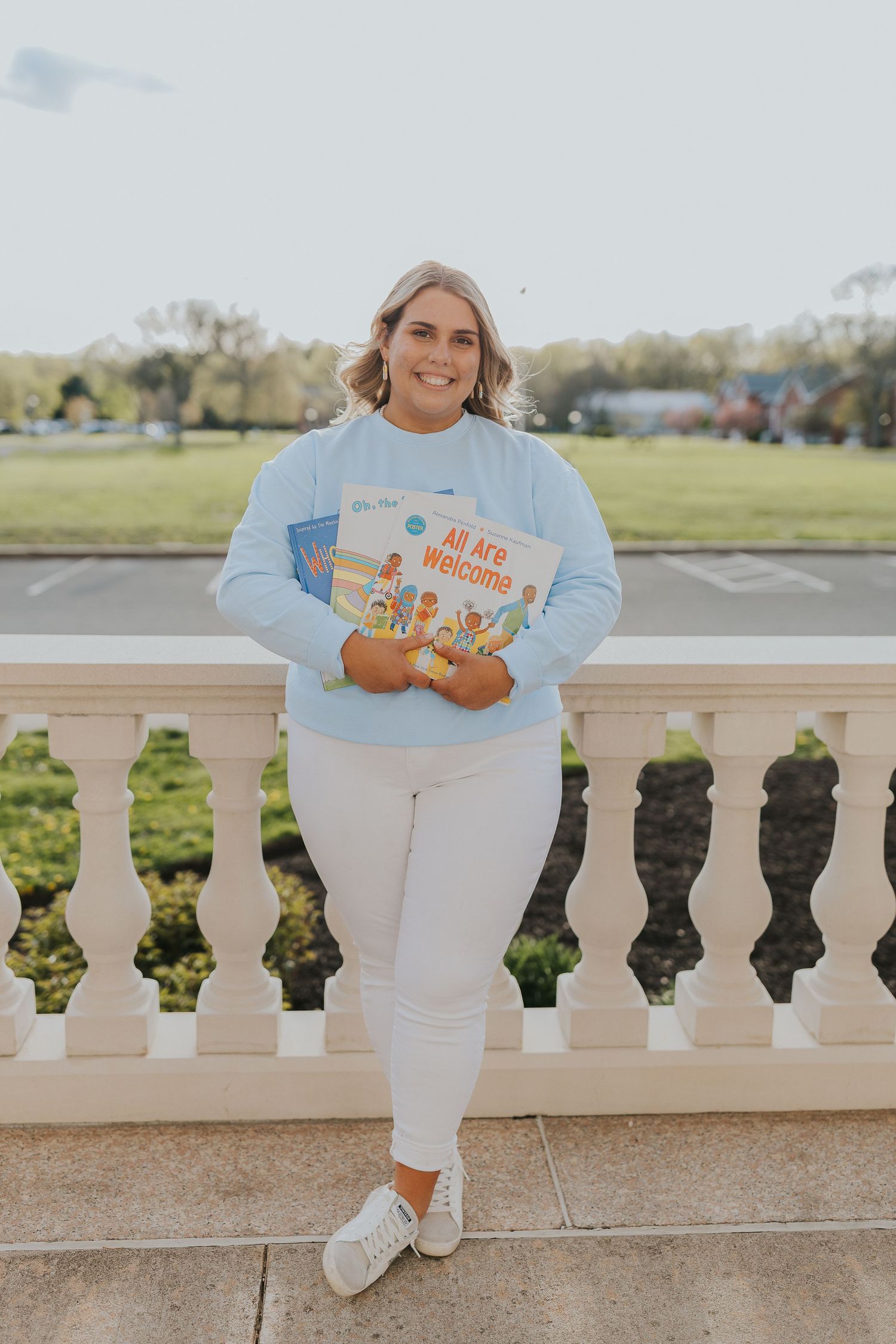 Smiling blonde woman in light blue sweatshirt holds childrens book on white balcony outdoors.