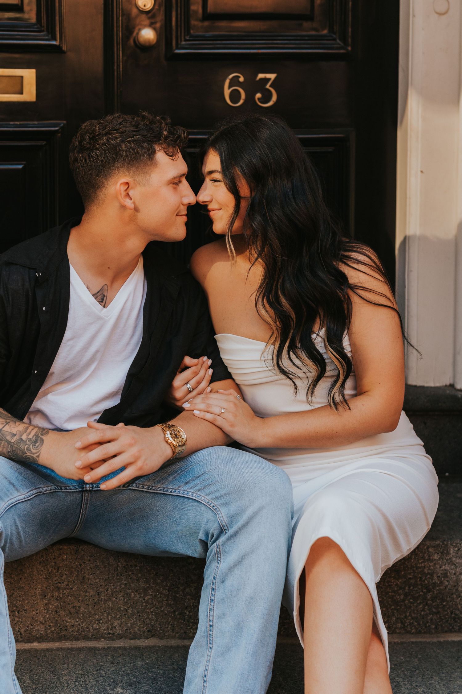 Romantic couple sitting on steps of a brownstone door numbered 63, leaning in for a kiss during engagement photos.