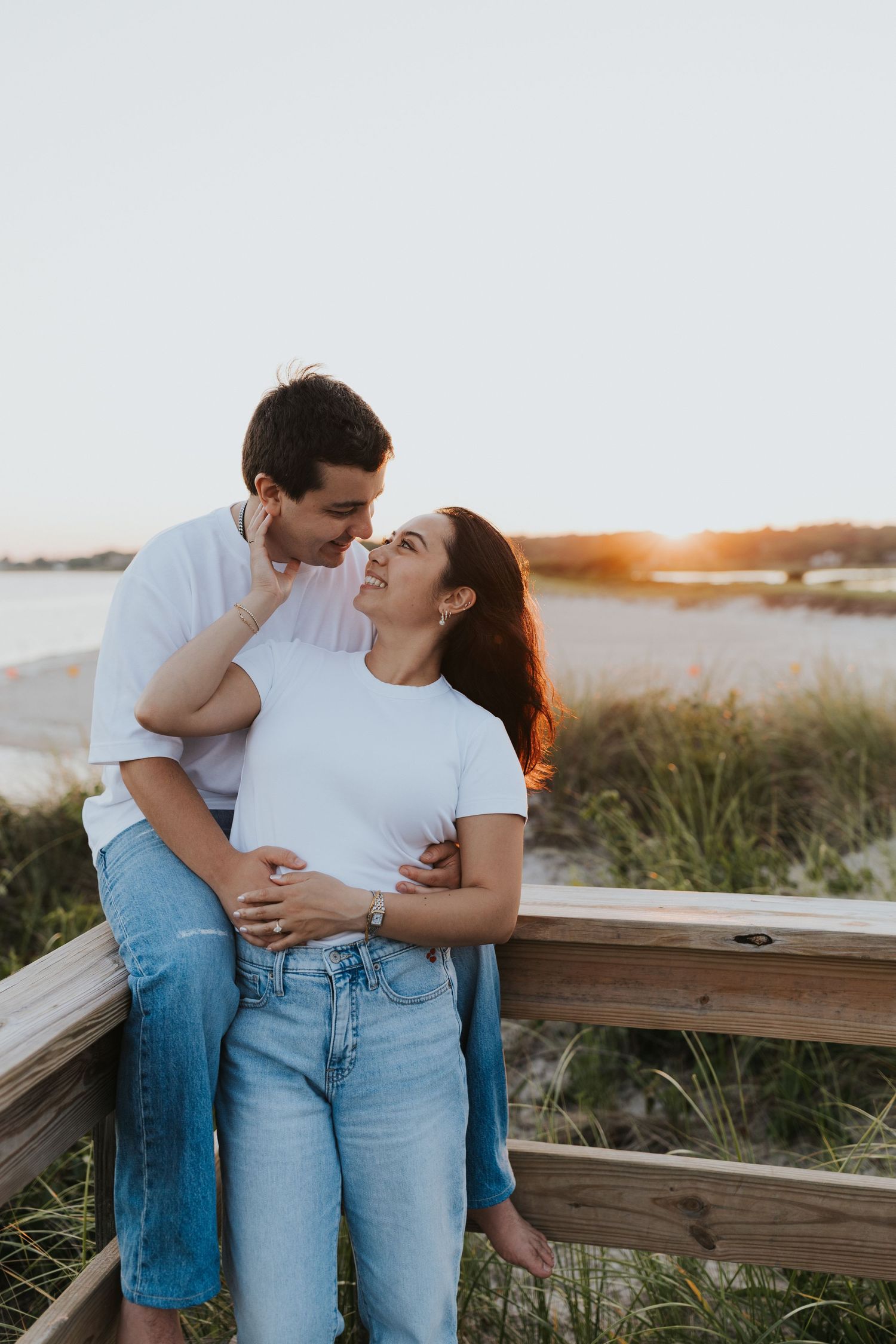 Couple sharing a romantic moment at sunset on a beach boardwalk, dressed in white tops and jeans.