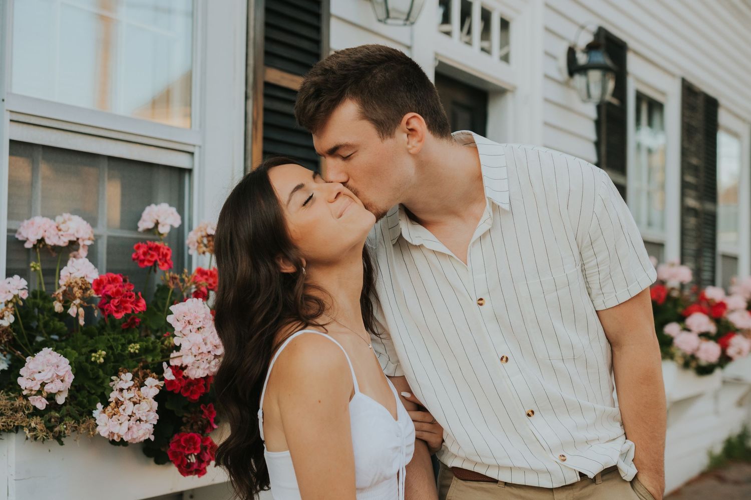 Couple sharing a romantic kiss in front of a white building adorned with red and pink flower boxes.