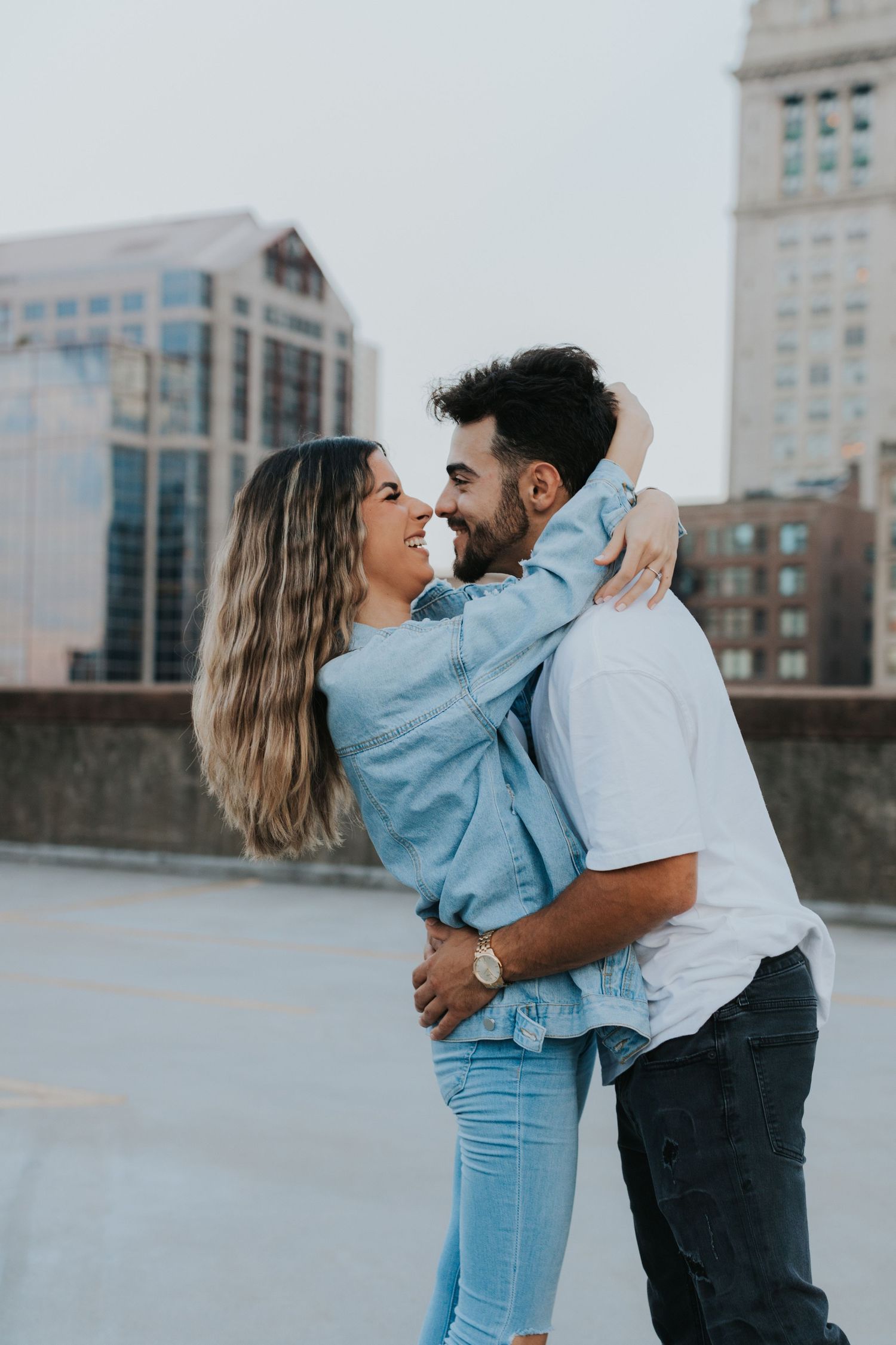 Happy couple embracing and smiling at each other on a rooftop with city buildings in the background.