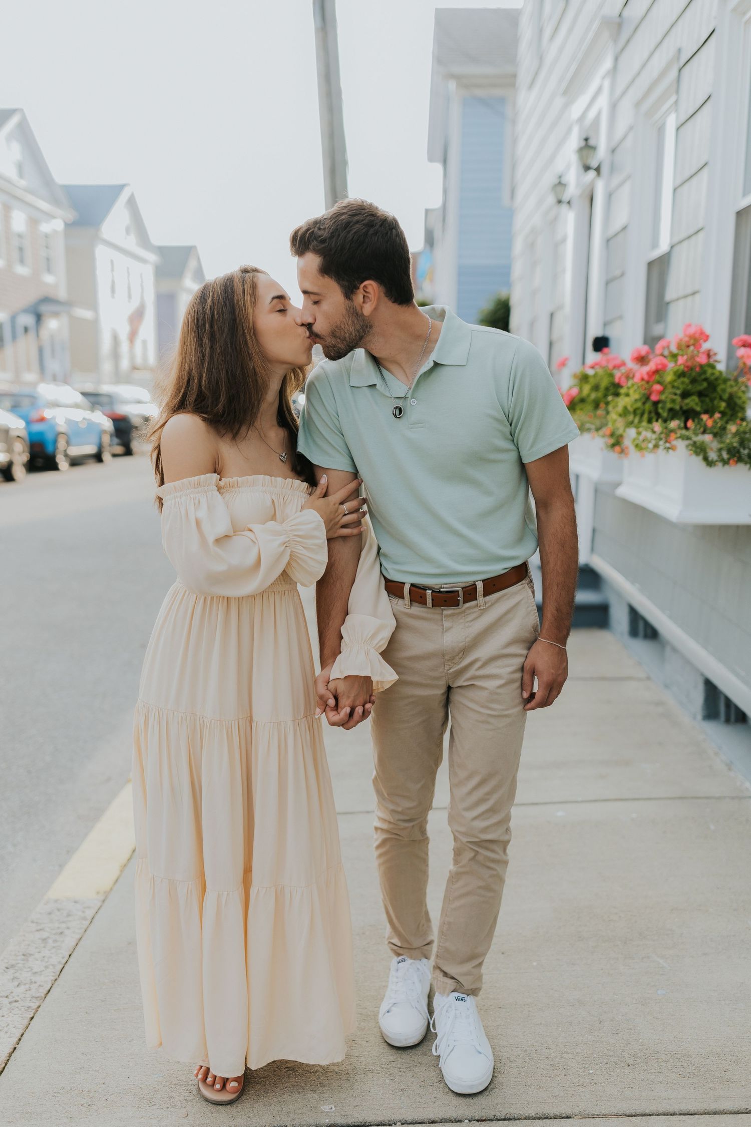 Couple sharing a kiss while holding hands on a charming sidewalk, woman in cream off-shoulder dress, man in sage polo.