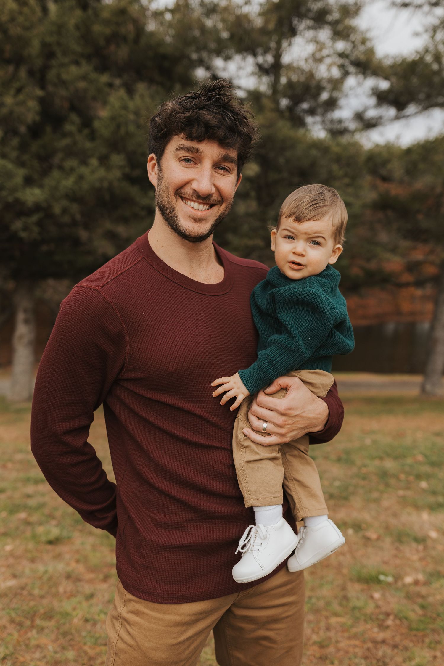 Smiling dark-haired father holding his baby boy in a park during autumn family portraits.