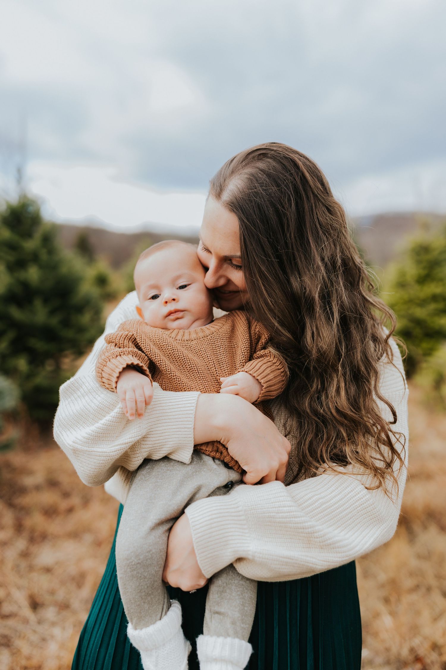 Mother with long wavy hair tenderly kisses baby in brown sweater at Christmas tree farm.