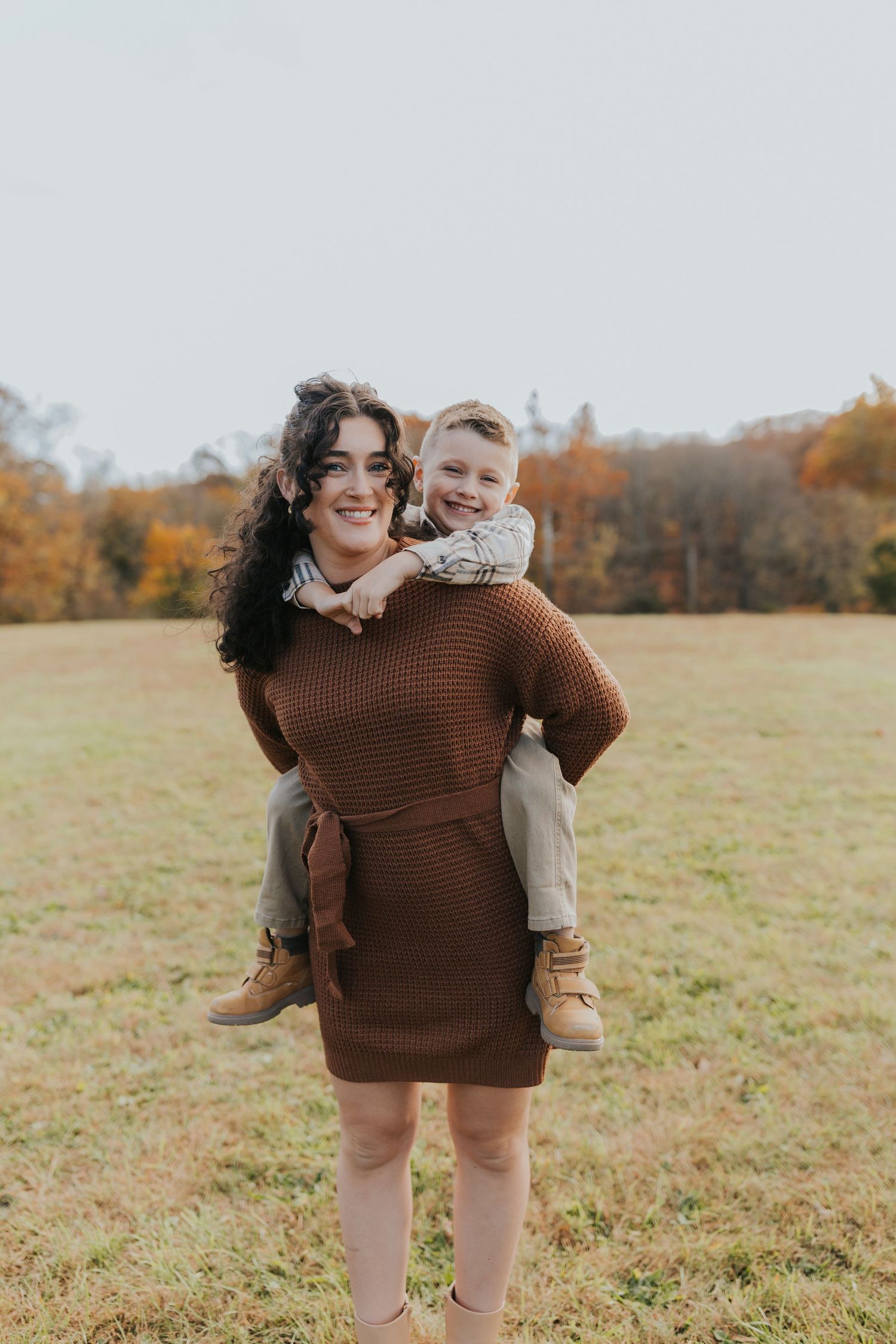 A smiling mother gives her young son a piggyback ride in an open grassy field during autumn.