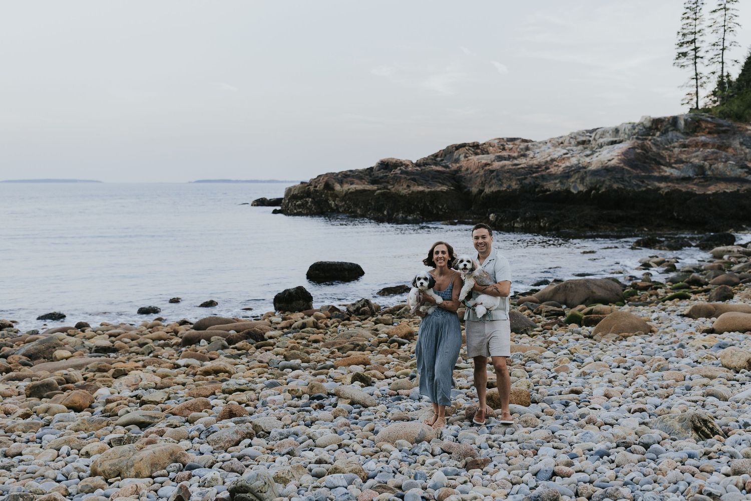 Couple standing on a rocky shoreline holding their dog with coastal cliffs behind.
