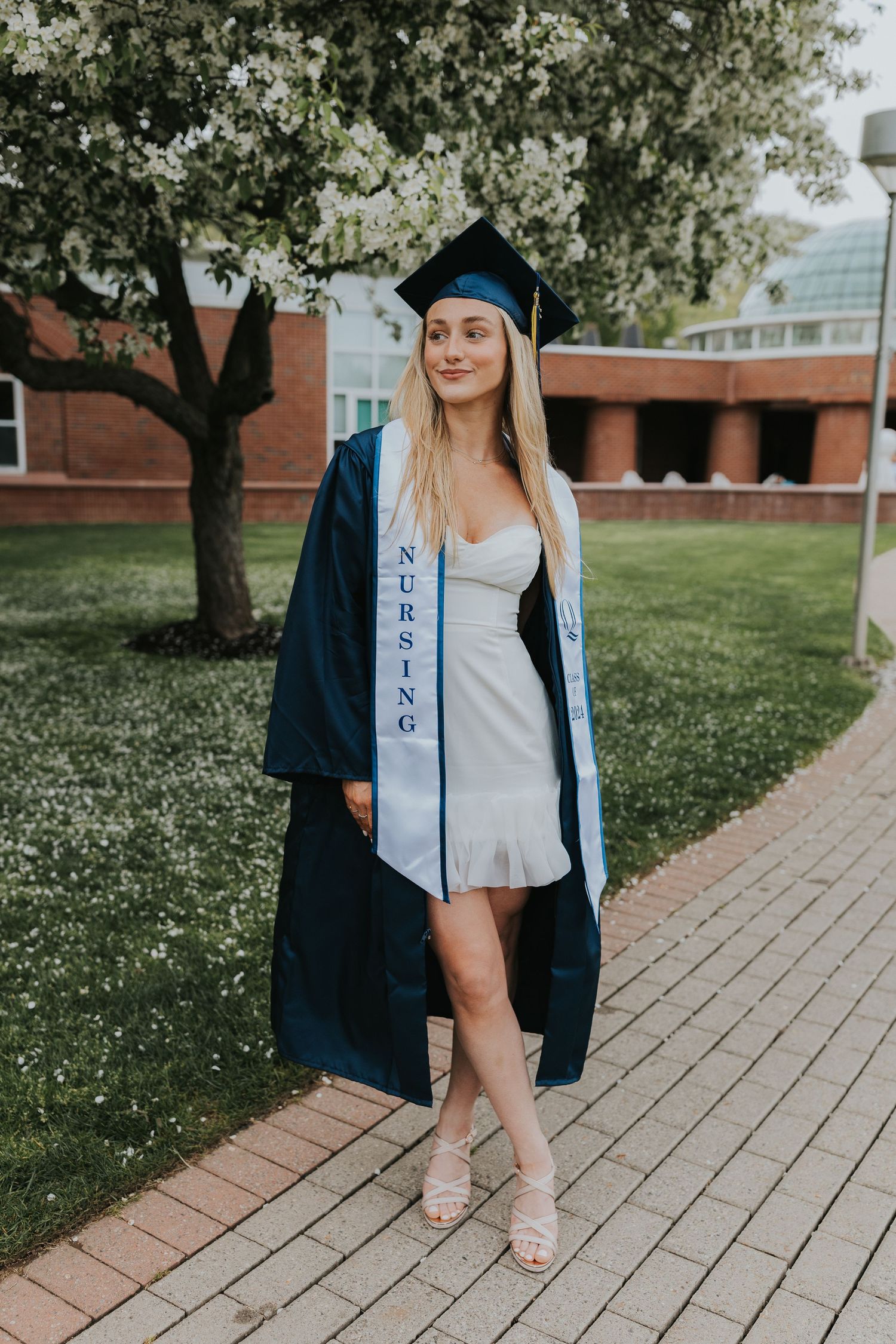 Nursing graduate posing in navy gown and white dress with graduation stole on a brick walkway.