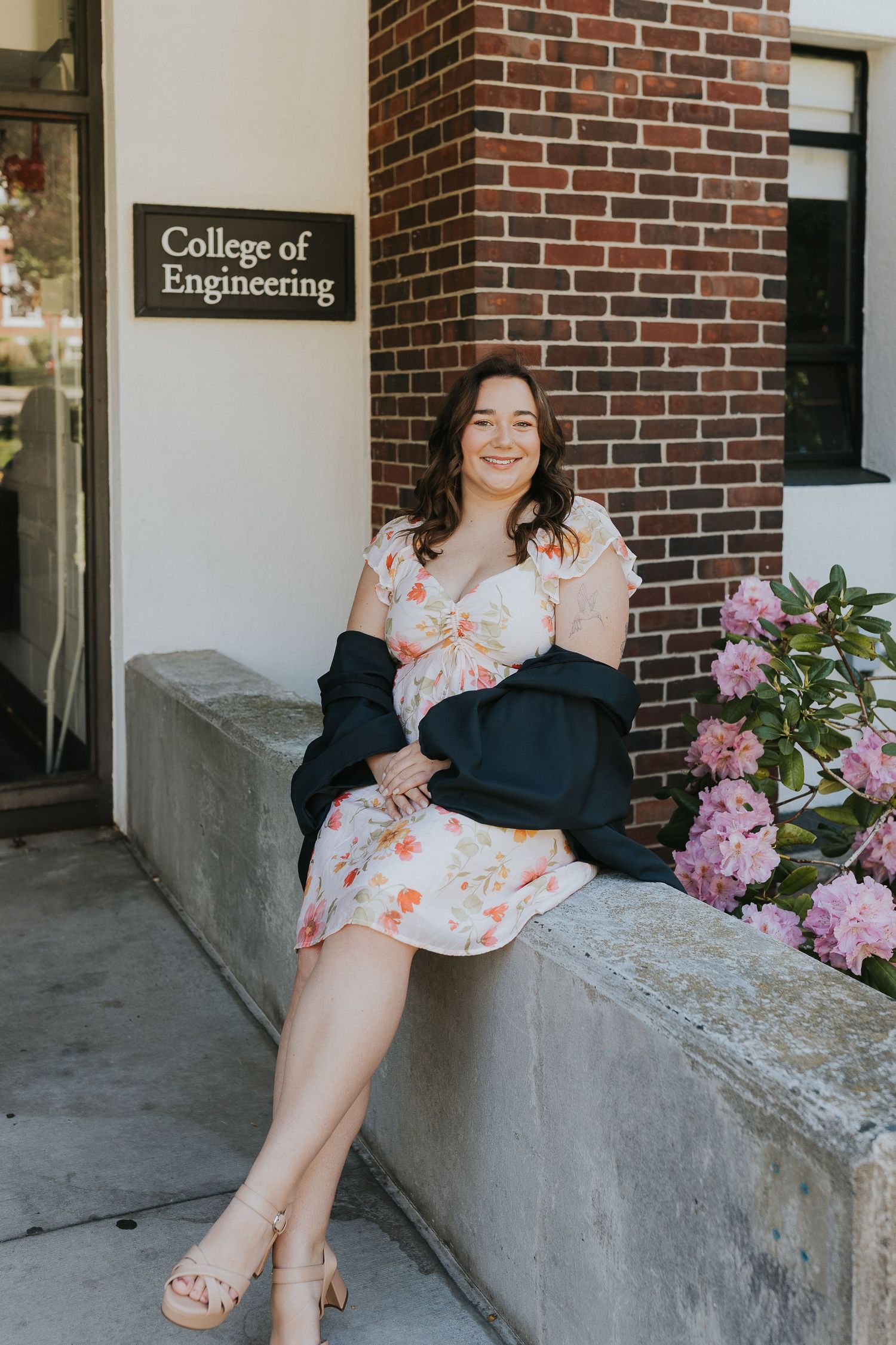 Woman in floral dress sits outside College of Engineering building with pink flowers.