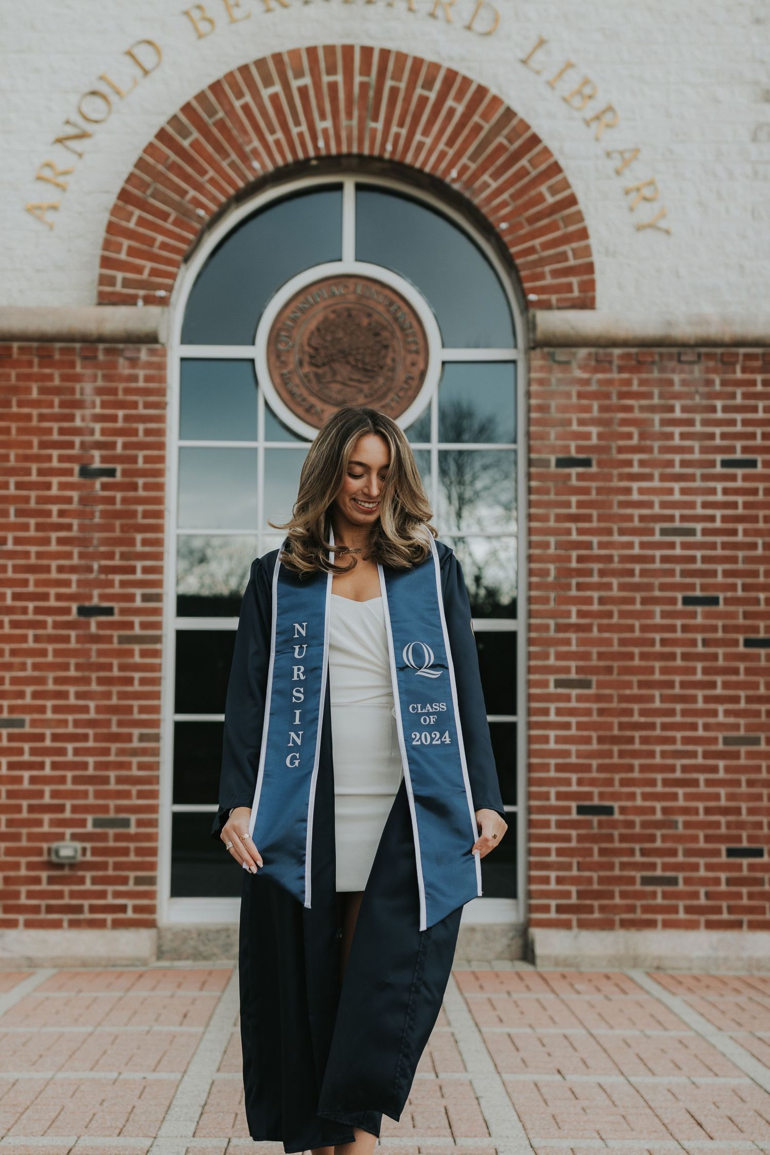 Nursing graduate in navy gown looks down at Class of 2024 stole outside Arnold Bernhard Library.