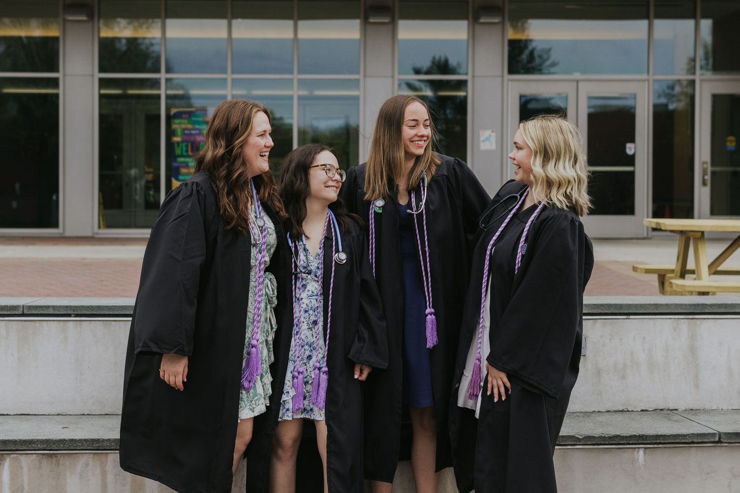 Four women in graduation gowns laugh together outside a university building.