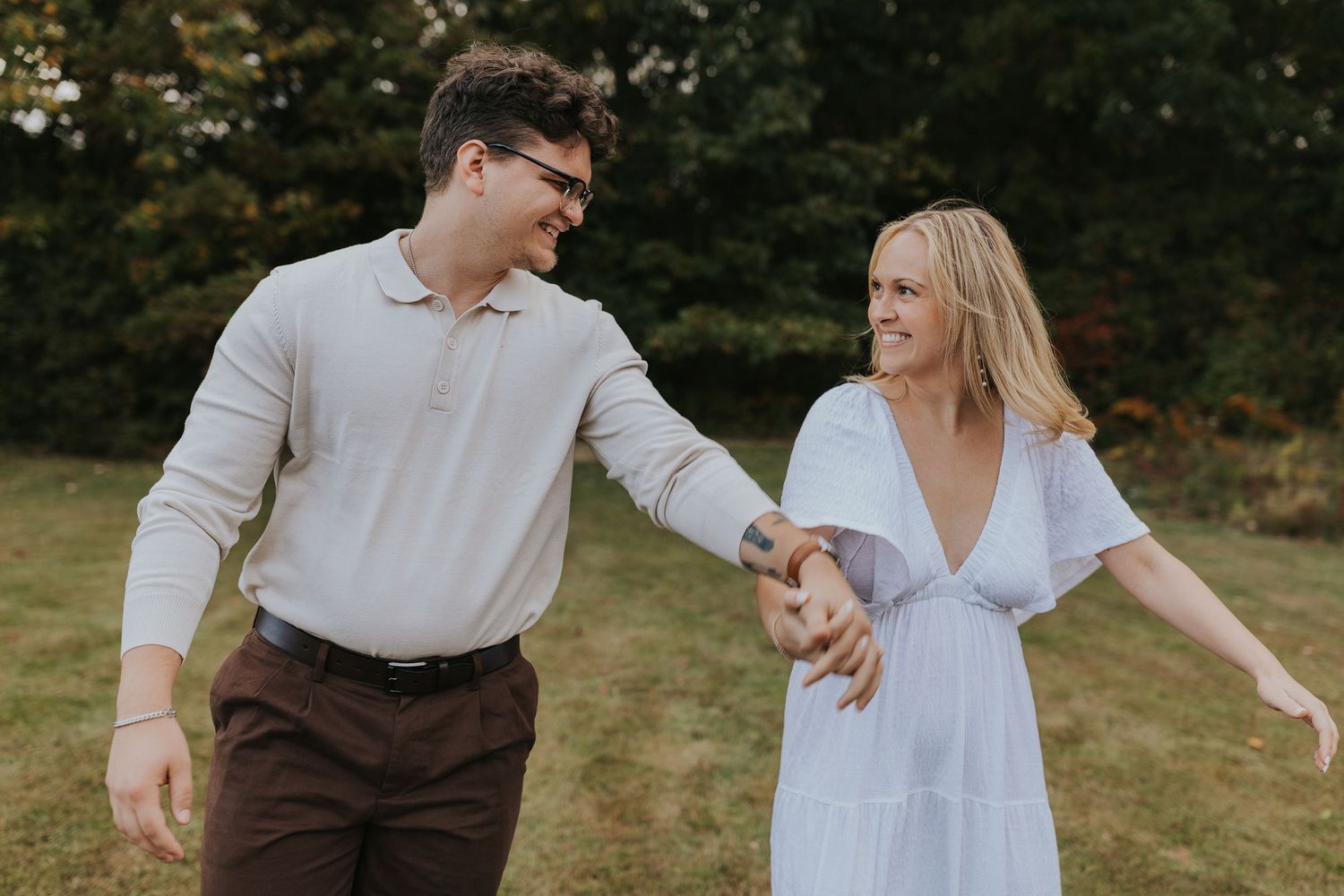 Happy couple walks hand in hand through a lush green park, smiling at each other in casual outfits.