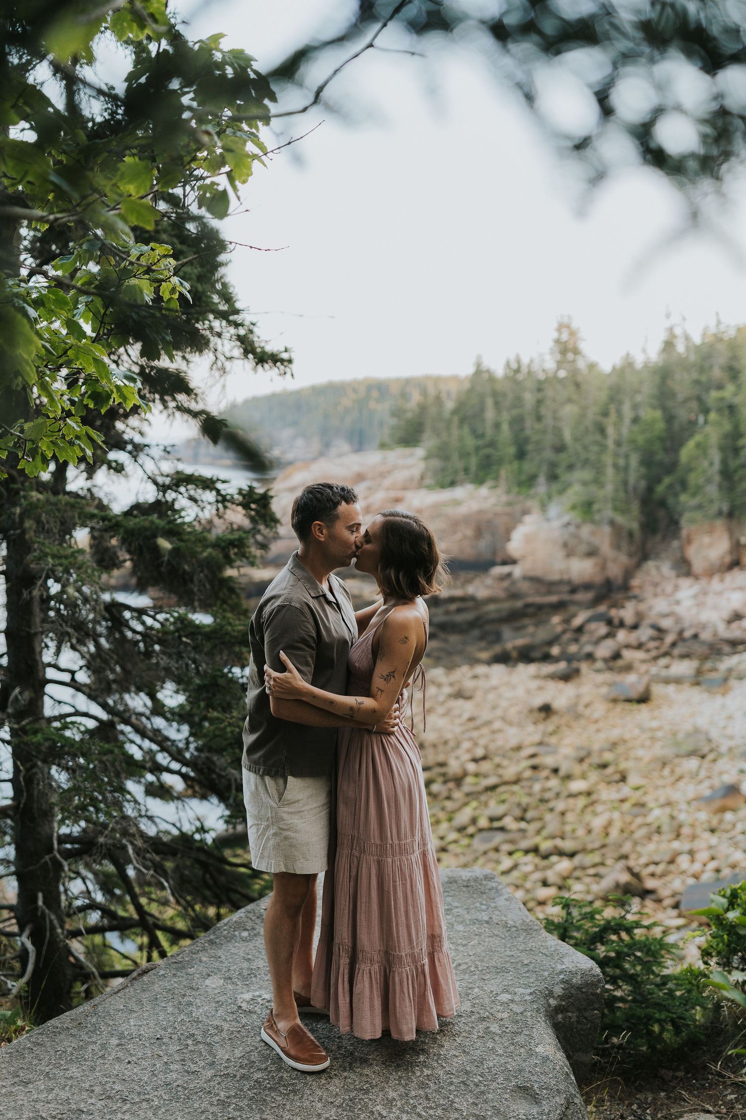 Couple sharing a romantic kiss on a rocky overlook surrounded by pine trees and coastal scenery.