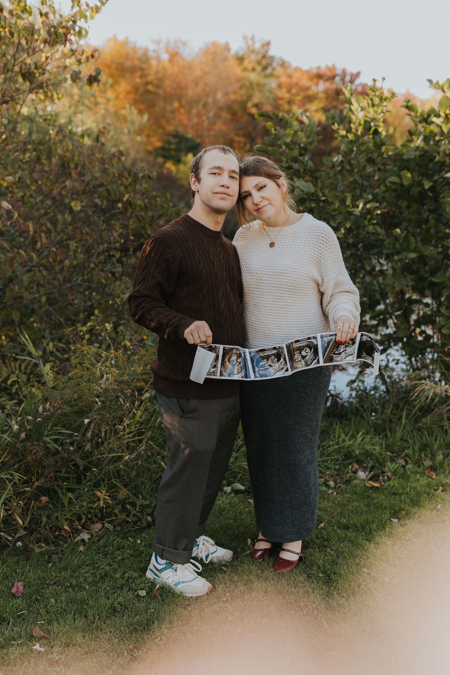 Expecting couple holding ultrasound photos outdoors during golden hour fall foliage pregnancy announcement session.