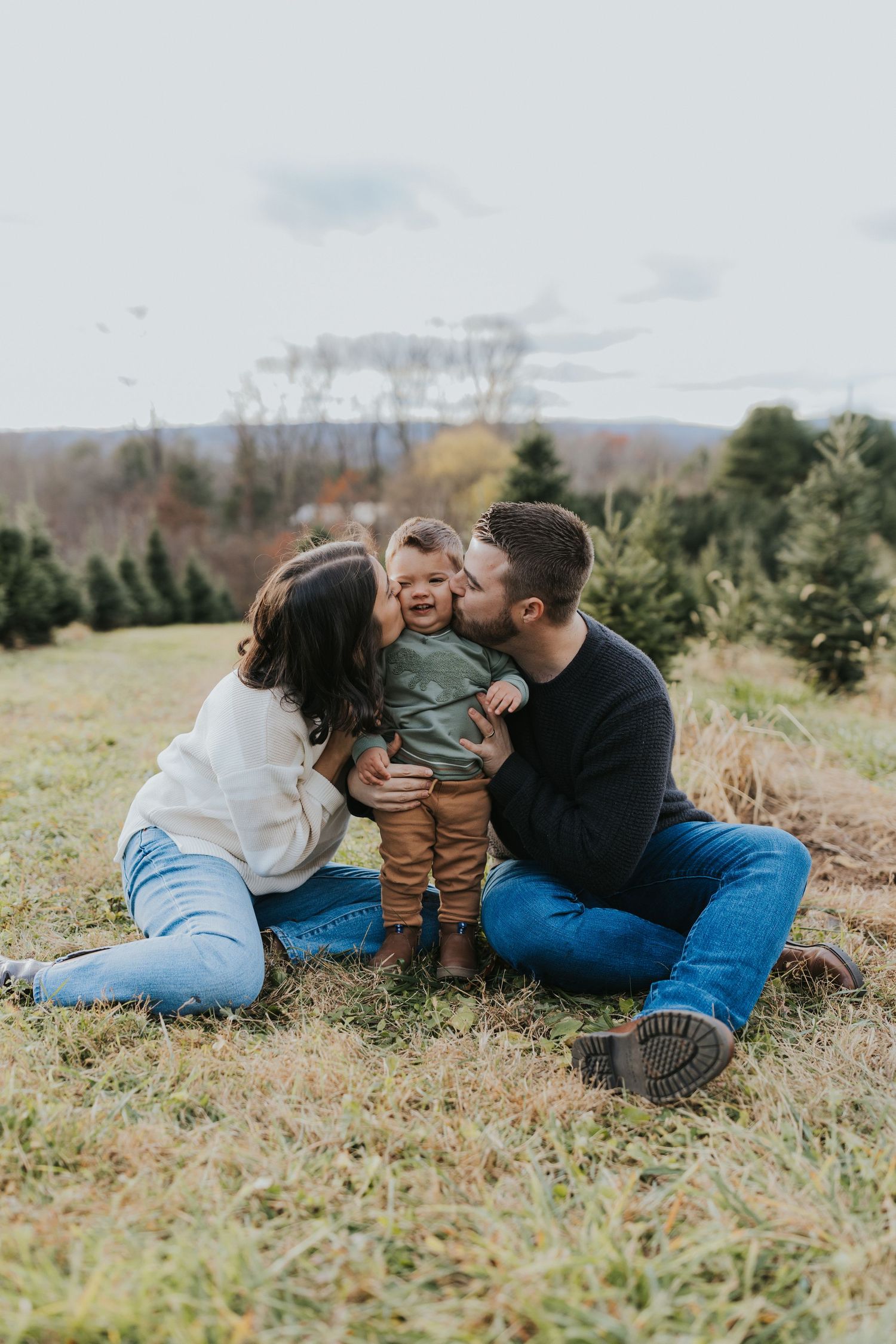 Two parents simultaneously kiss their happy toddler on the cheeks while sitting at a Christmas tree farm.