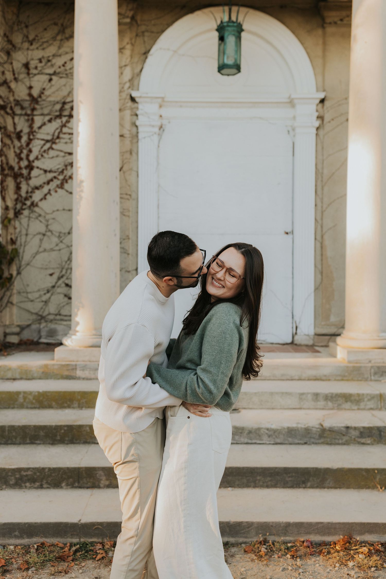 Couple embracing and laughing on steps of white columned building, man kissing woman's cheek in playful moment.