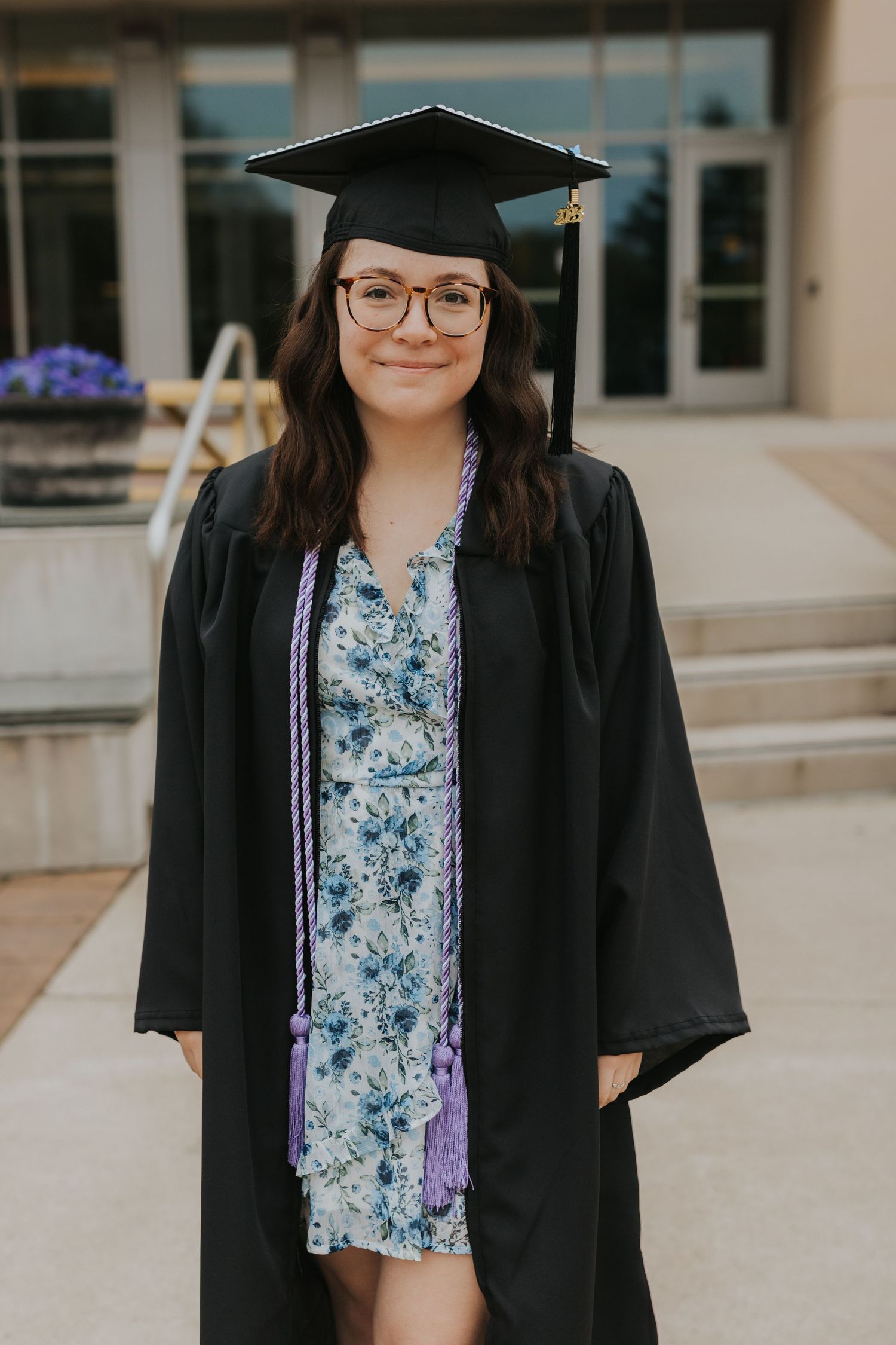Female graduate smiling in black cap and gown with purple honor cords outside university building in floral dress.