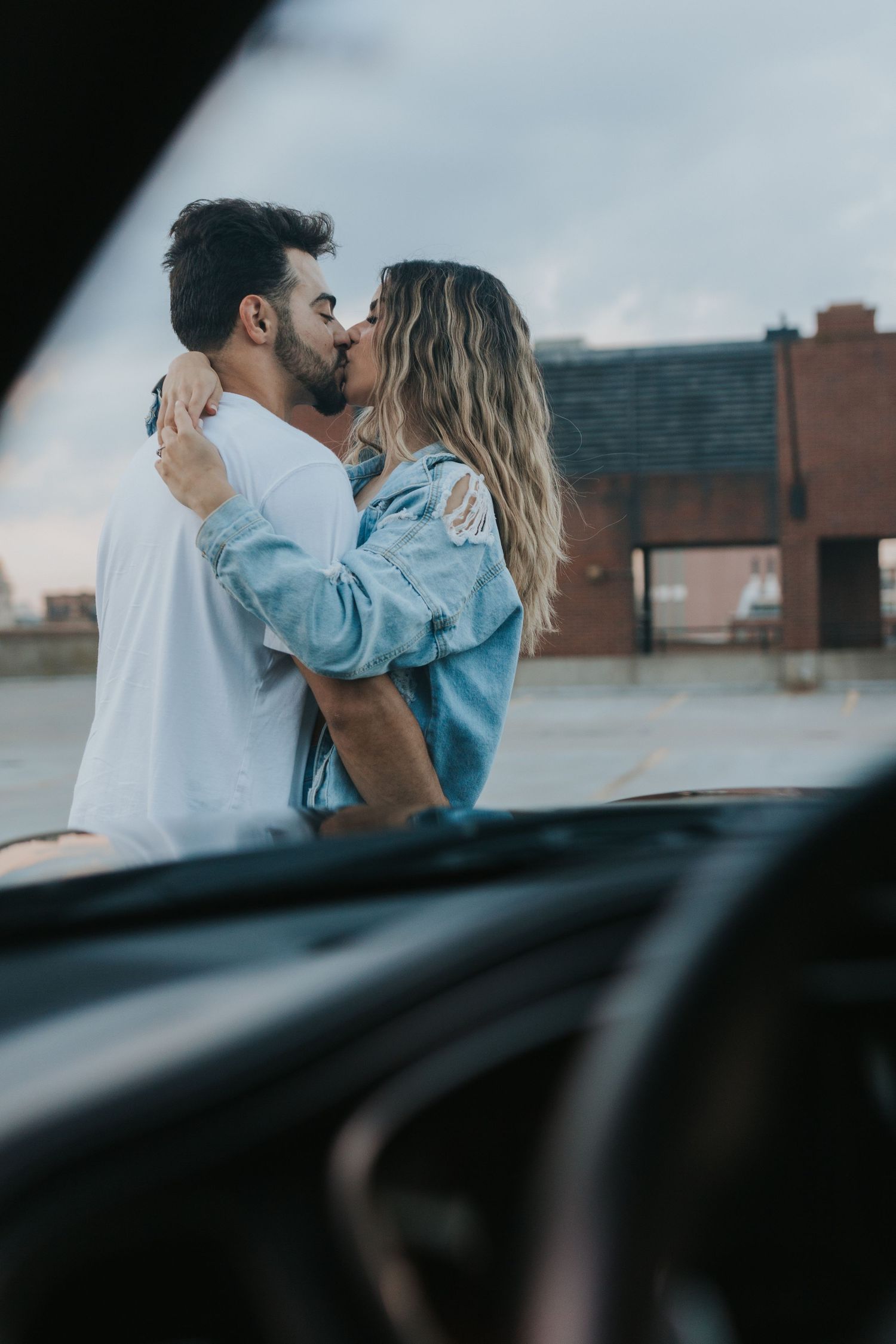 Couple sharing a romantic kiss outdoors near a car on a rooftop parking lot under cloudy skies.
