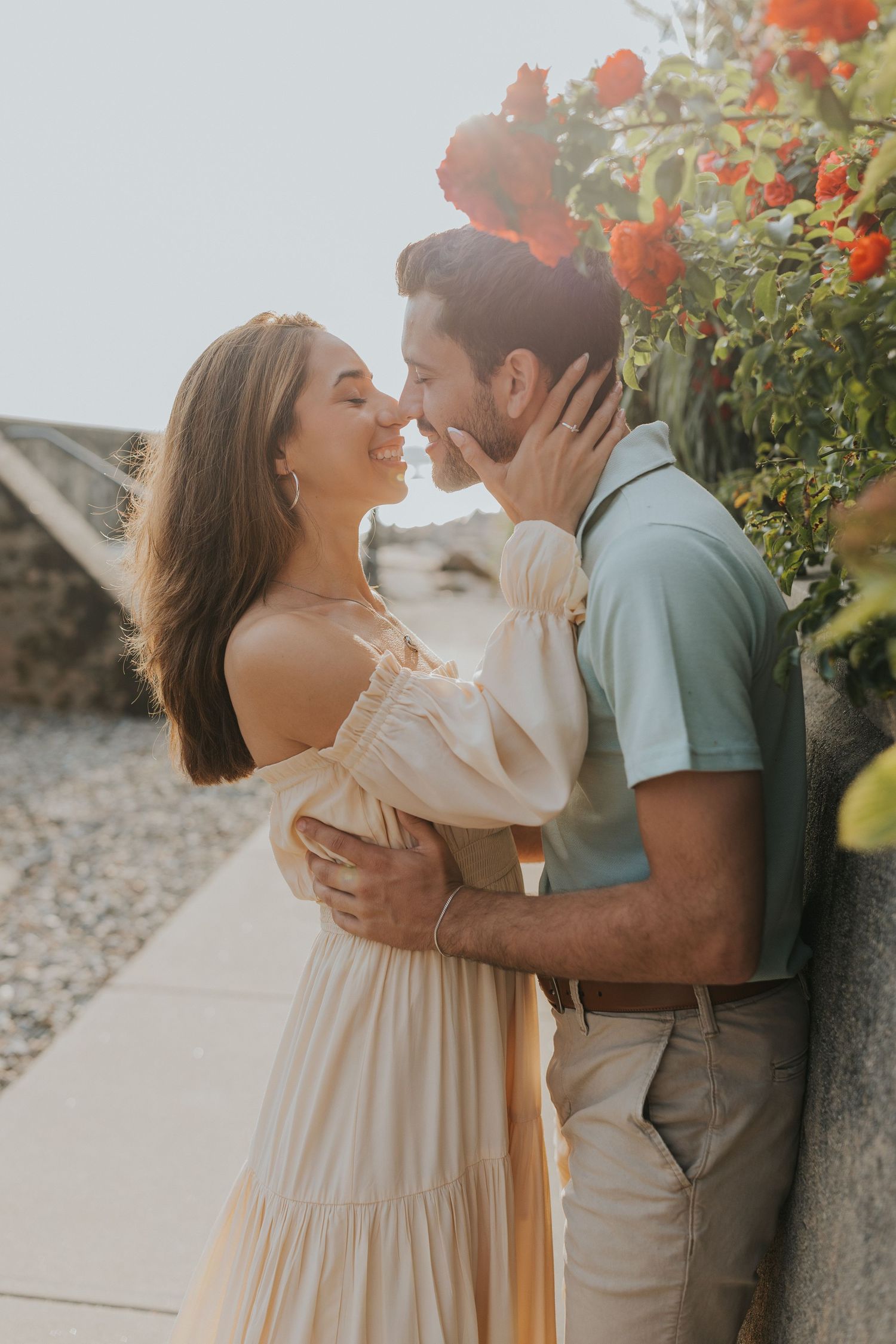 Couple sharing a romantic almost-kiss against stone wall with red roses, bathed in warm golden sunset light.