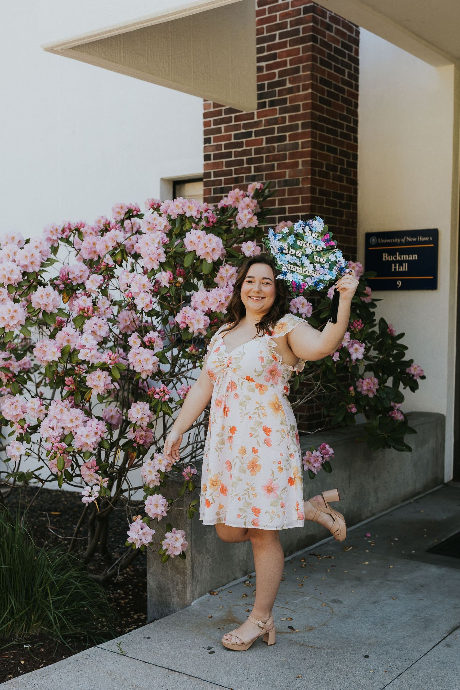 Joyful graduate in floral dress holding colorful bouquet beside blooming pink rhododendrons at Buckman Hall.